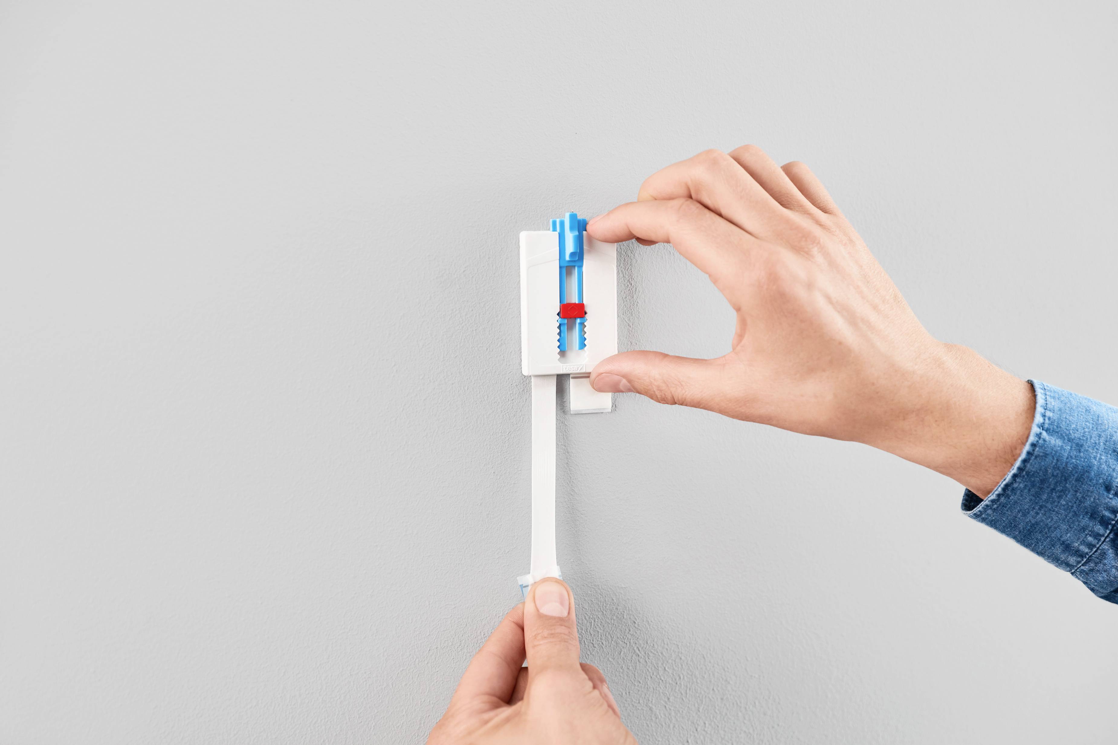 Osoba mocuje niebiesko-czerwoną kołkę rozporową do szarej ściany za pomocą rąk.