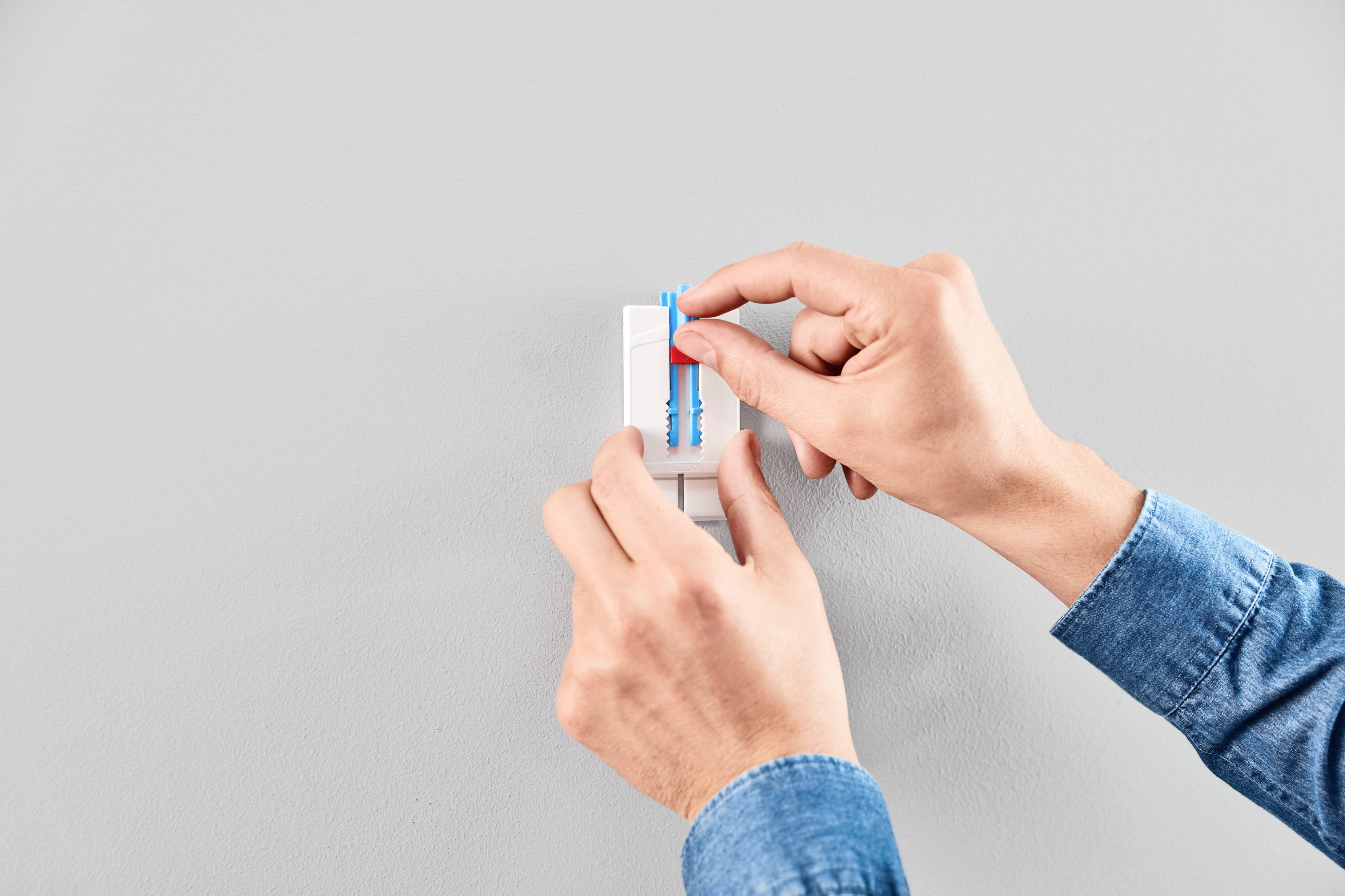 Dłonie osoby włączającej przełącznik światła na szarej ścianie. Osoba ma na sobie niebieską koszulę.