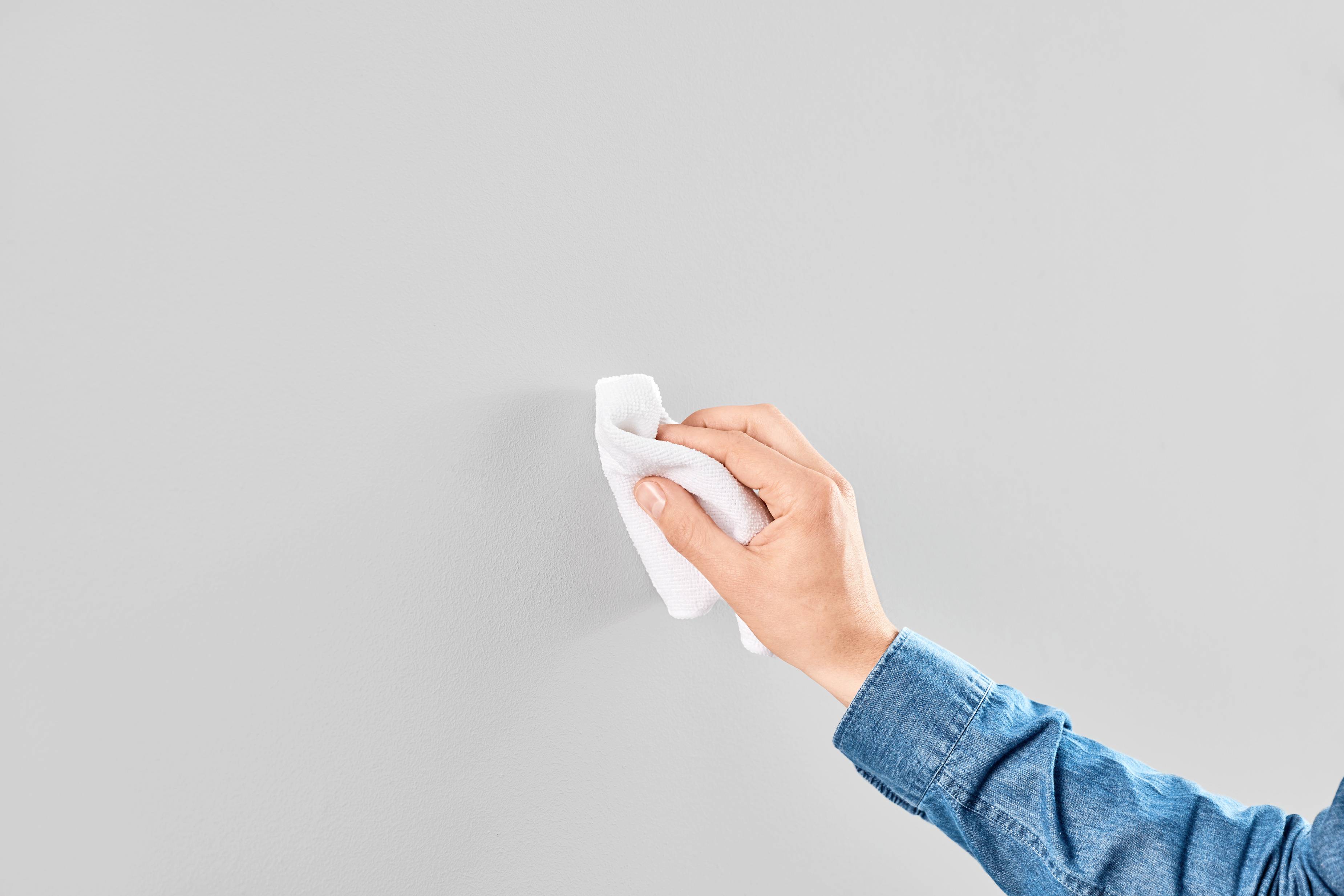 Ręka przesuwa białą ścierką po szarej ścianie, czyścząc ją. Osoba ma na sobie niebieską koszulę.