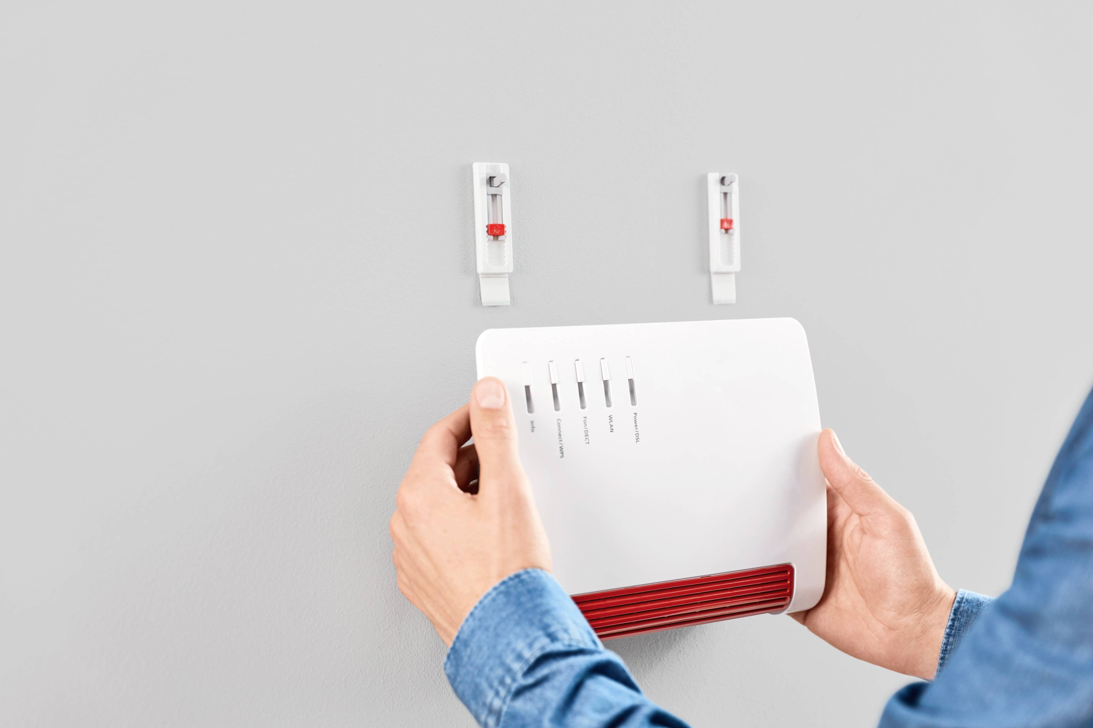 Osoba mocuje biały router Wi-Fi na ścianie. Router wyświetla symbole siły sygnału i połączenia. Widoczne są dwa puste haczyki.