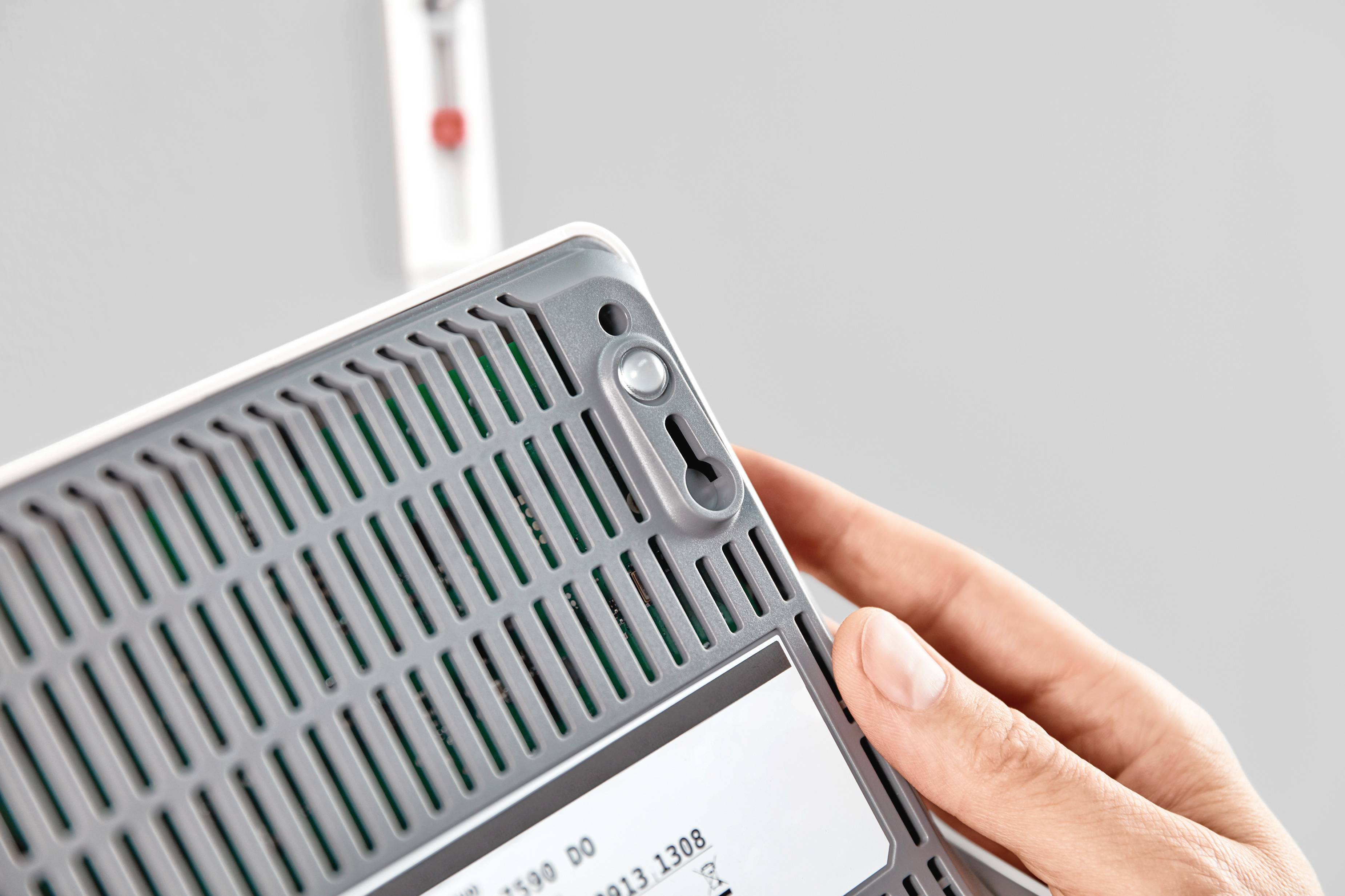 Osoba trzyma szary przedmiot z otworami wentylacyjnymi i białą etykietą. Tło rozmyte z czerwoną plamką.