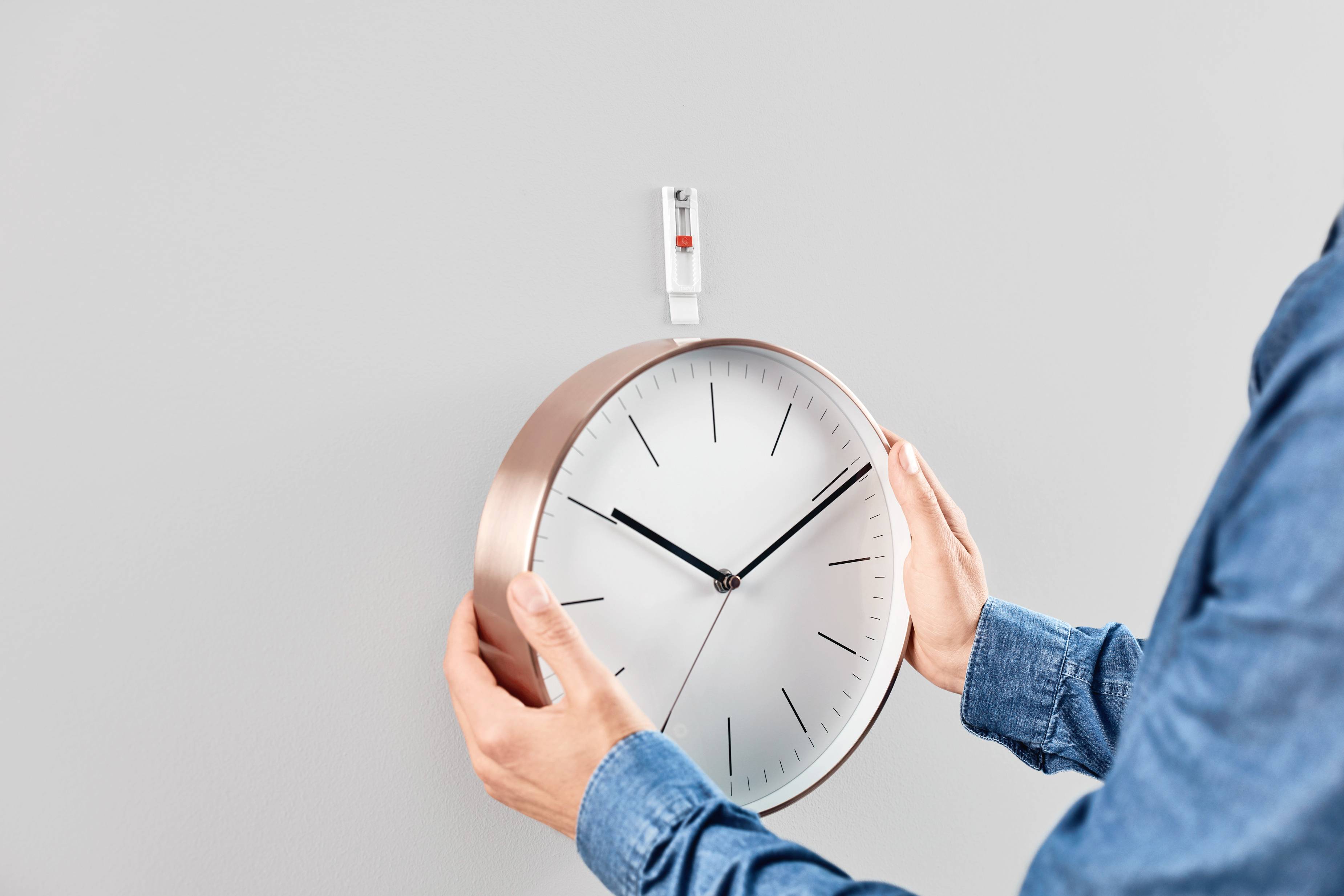 Osoba zawiesza okrągły zegar ścienny z miedzianą ramą na haczyku na ścianie. Zegar wskazuje godzinę 10:10.
