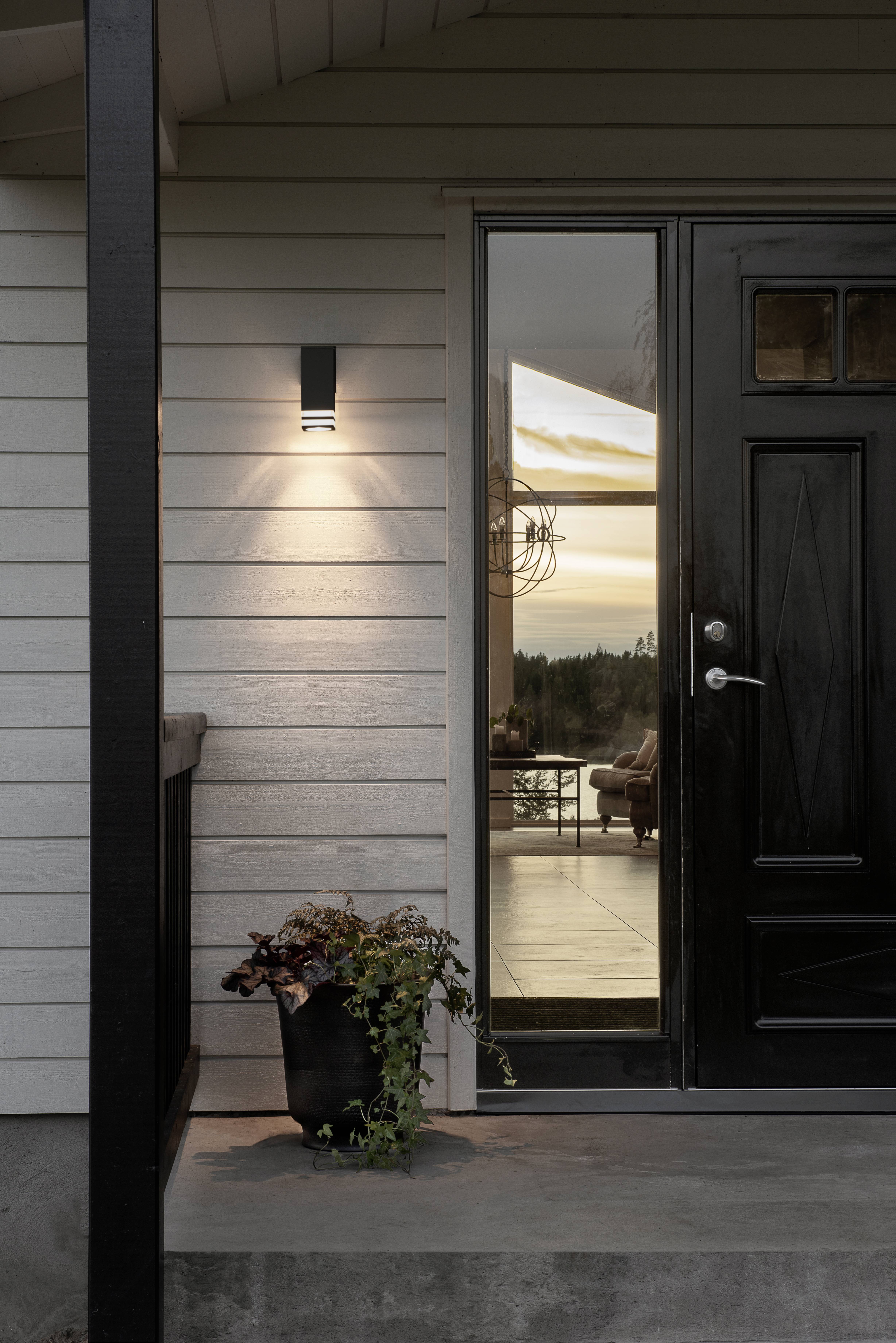 Nowoczesna weranda o zachodzie słońca, z oświetloną lampą zewnętrzną, czarnymi drzwiami i oknem, przez które widać przytulne wnętrze.