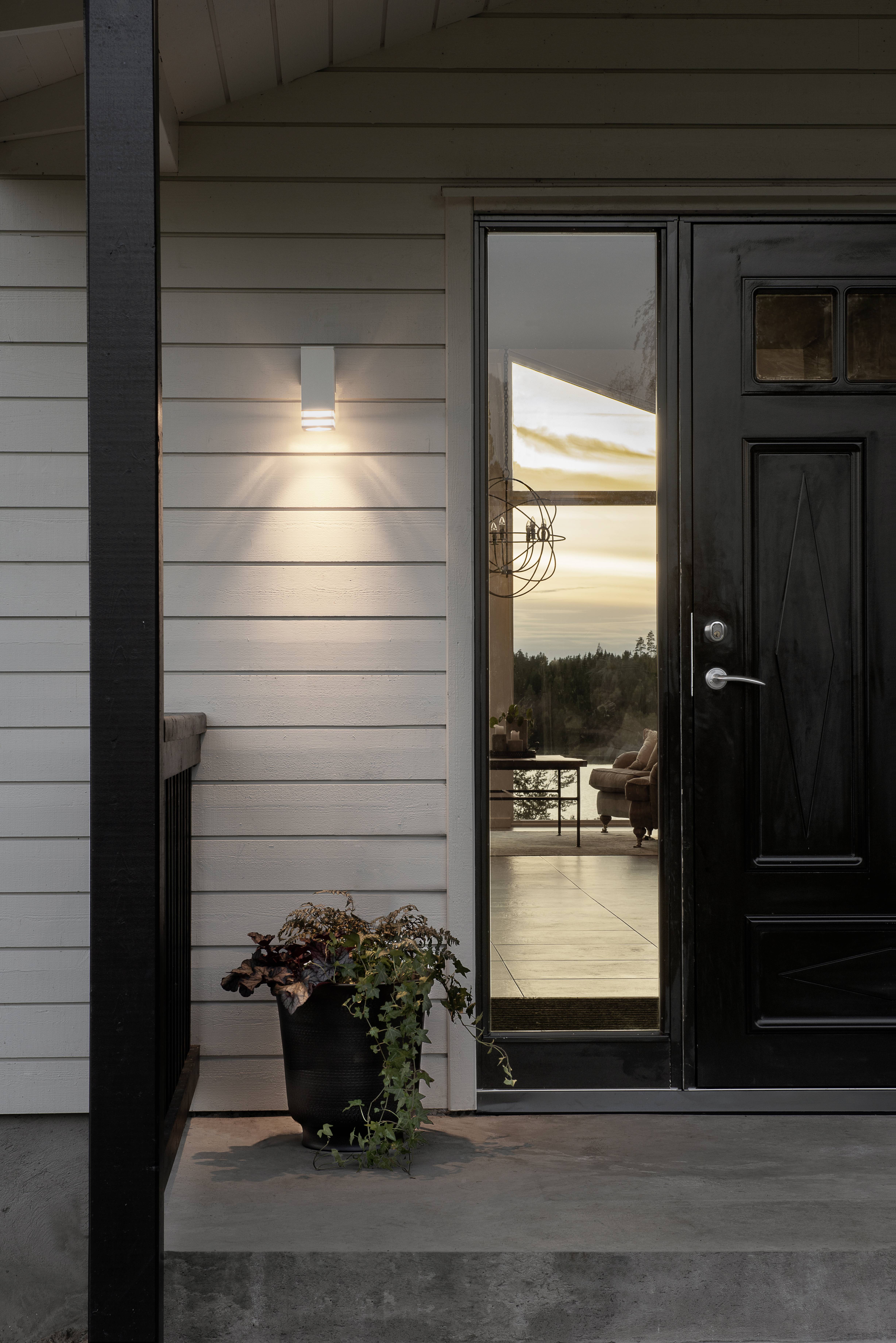 Drzwi wejściowe i przeszklona ściana nowoczesnego domu, oświetlone lampą外外外外ą. W tle zachodzące słońce i drzewa.