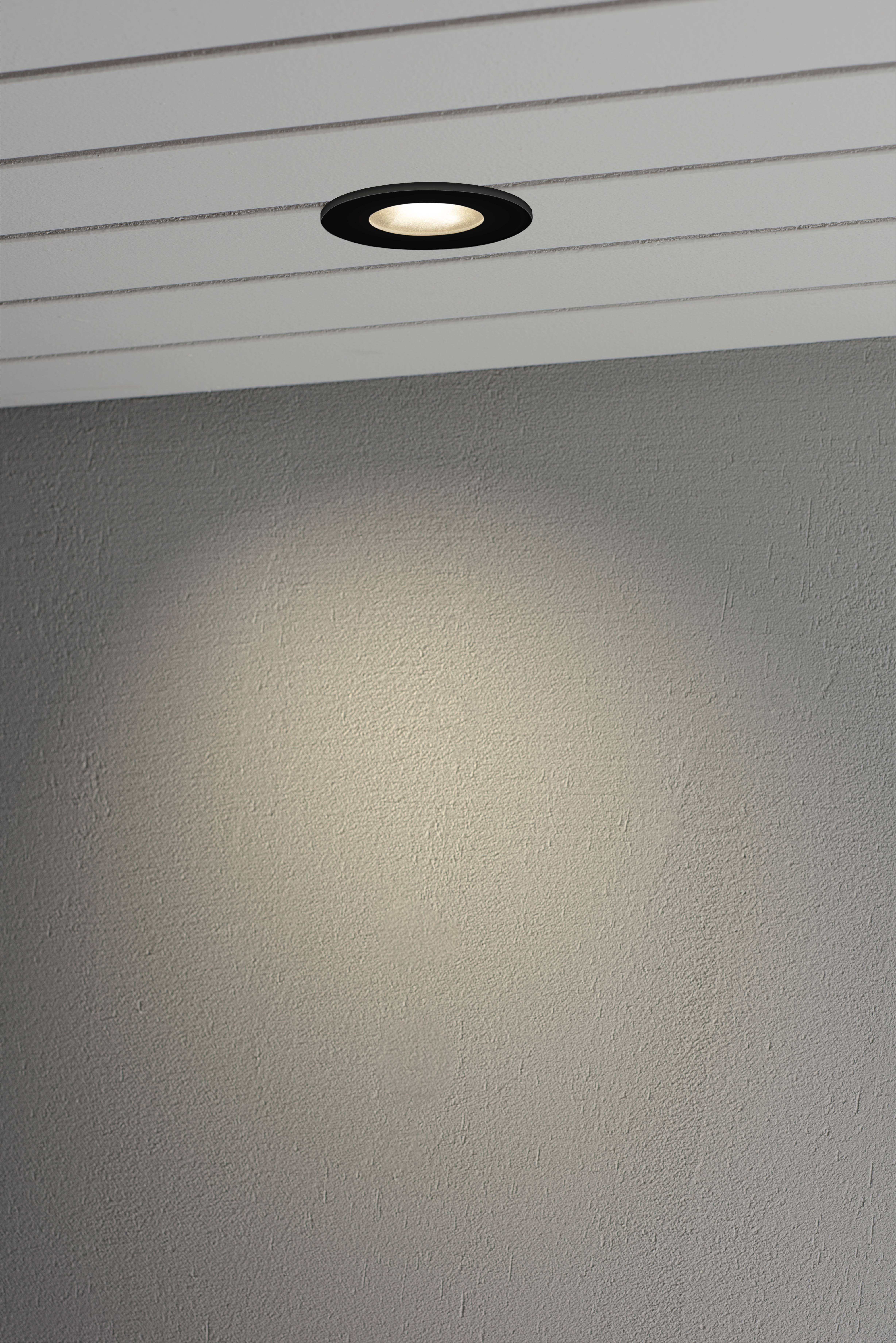 Wpuszczany reflektor sufitowy w czarnej obudowie, rzuca skoncentrowane światło na szarą ścianę, tworząc świetlny okrąg.