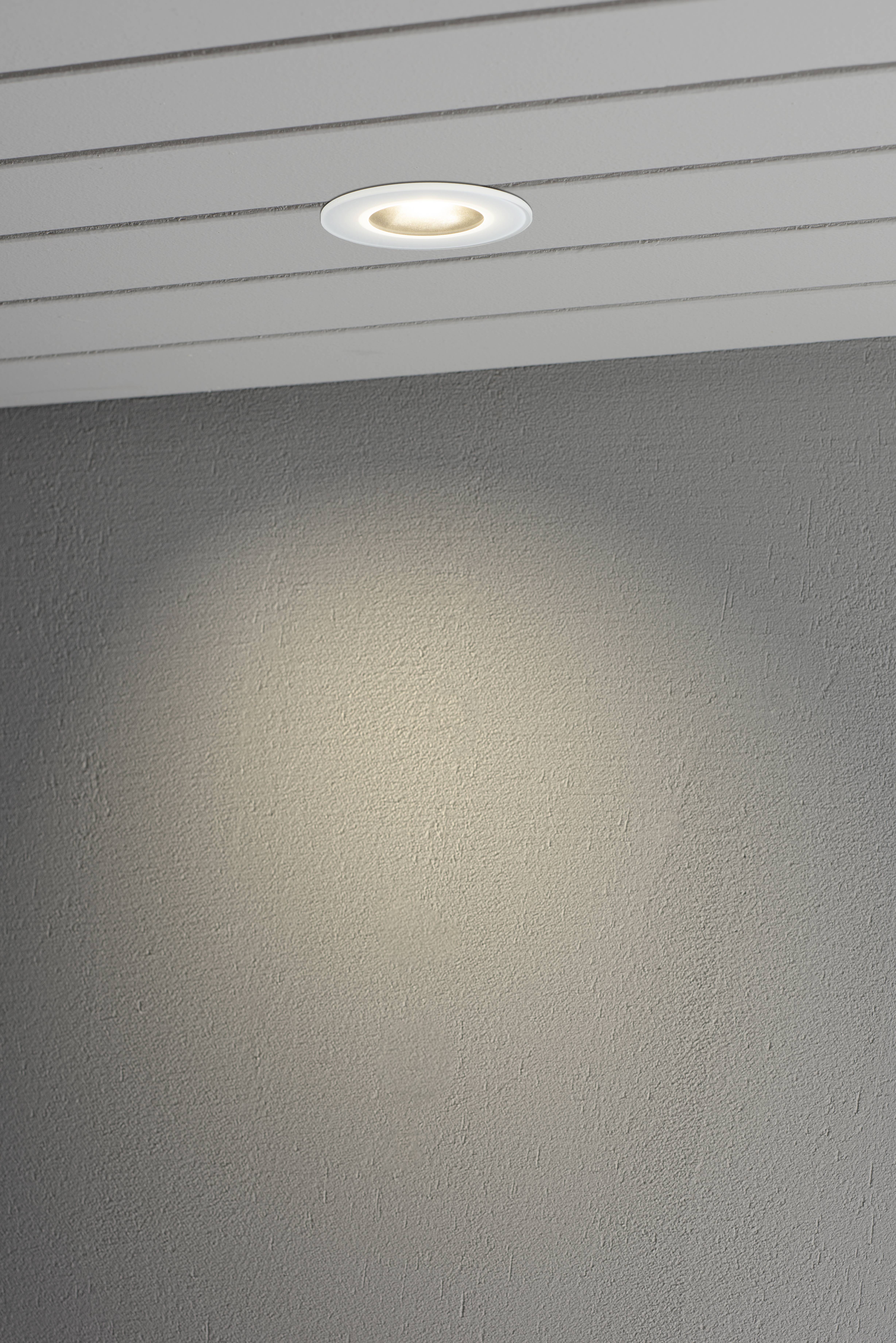 Oprawa sufitowa rzuca miękkie pasmo światła na szarą ścianę. Nowoczesne wnętrze z precyzyjnym oświetleniem.