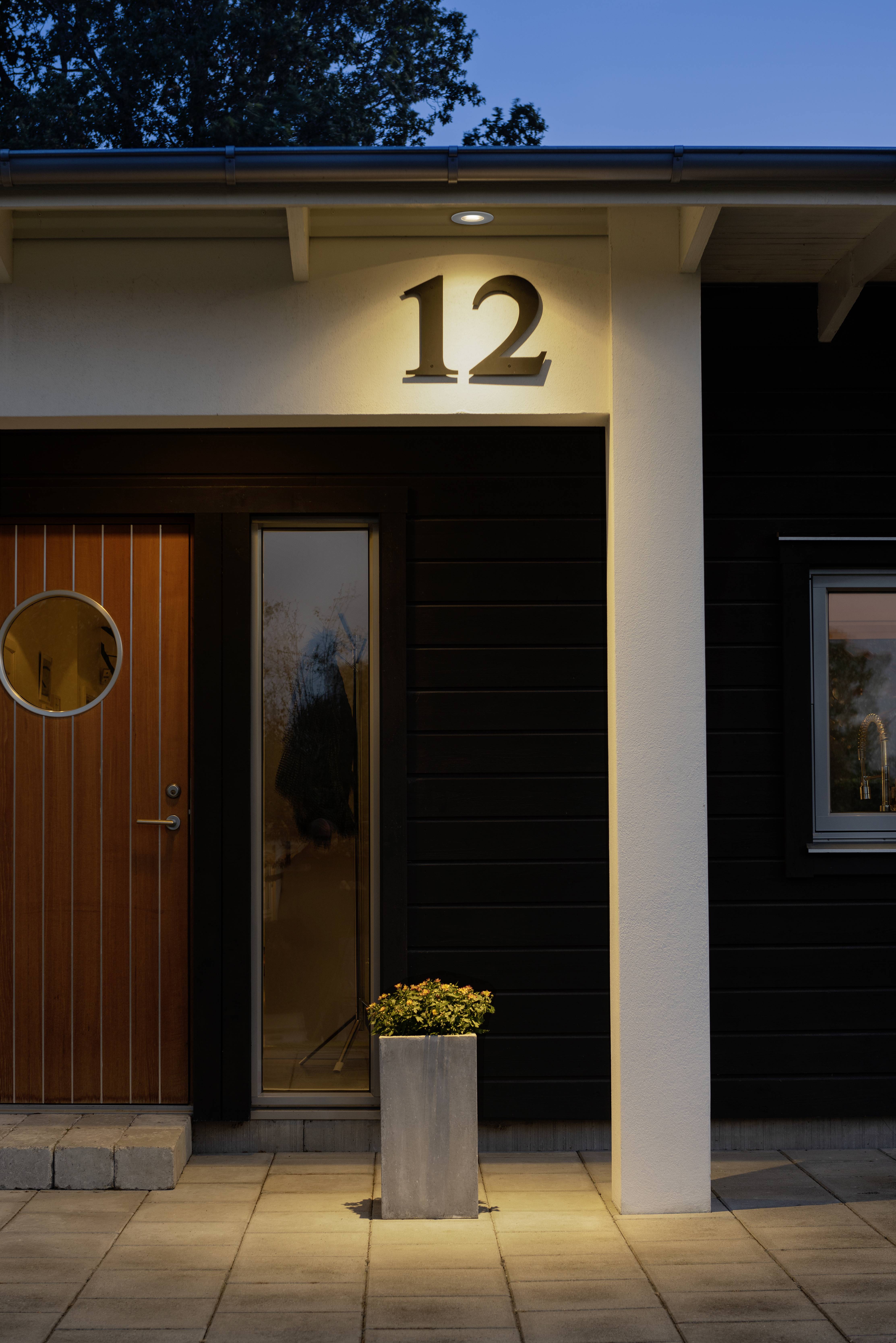 Wejście nowoczesnego domu o zmierzchu, oświetlone z numerem domu 12 nad drzwiami, doniczka z kwiatami na pierwszym planie.