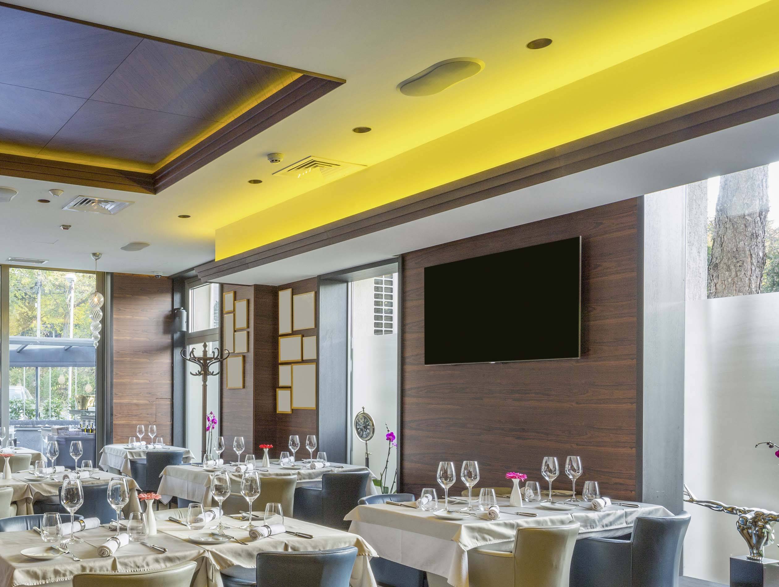 Elegancka restauracja z nakrytymi stołami, białymi obrusami i kieliszkami do wina. Ściany są wyłożone drewnem, a miękkie światło sufitowe tworzy przytulną atmosferę.