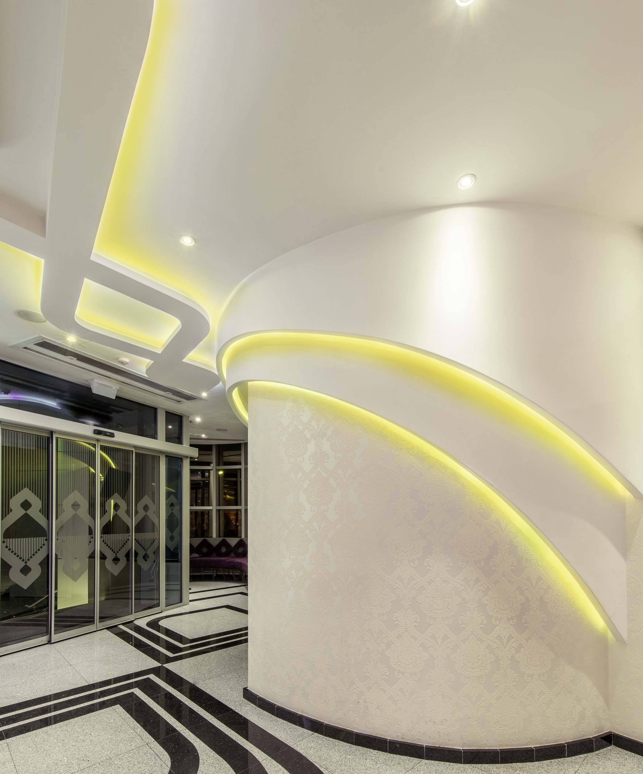 Nowoczesne foyer z zakrzywionymi ścianami i żółtymi lampami LED. Czarno-biały geometryczny wzór podłogi oraz szklane drzwi po lewej stronie.