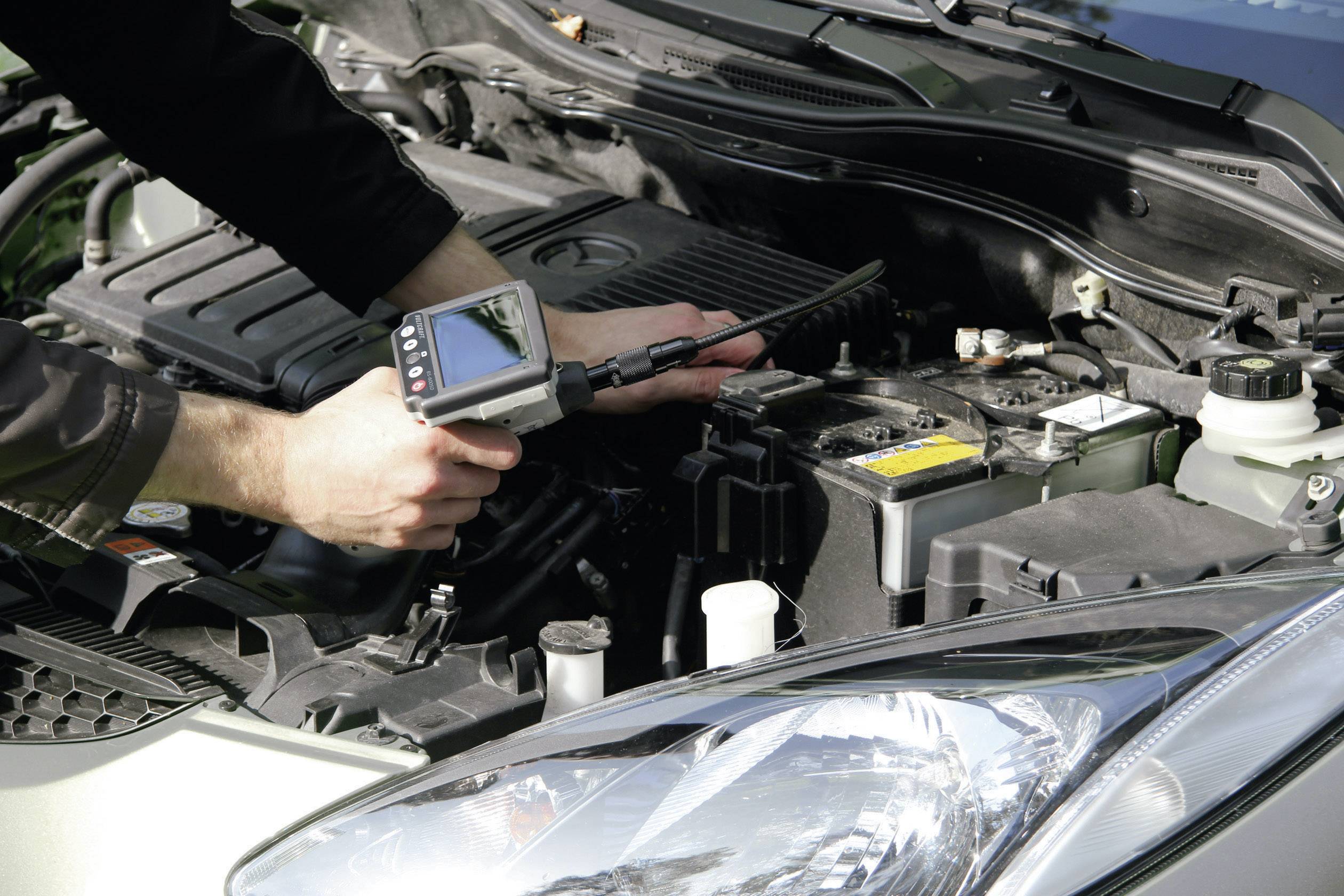 Osoba używa urządzenia diagnostycznego do sprawdzenia silnika samochodu. Komora silnika jest otwarta i dobrze widoczna.