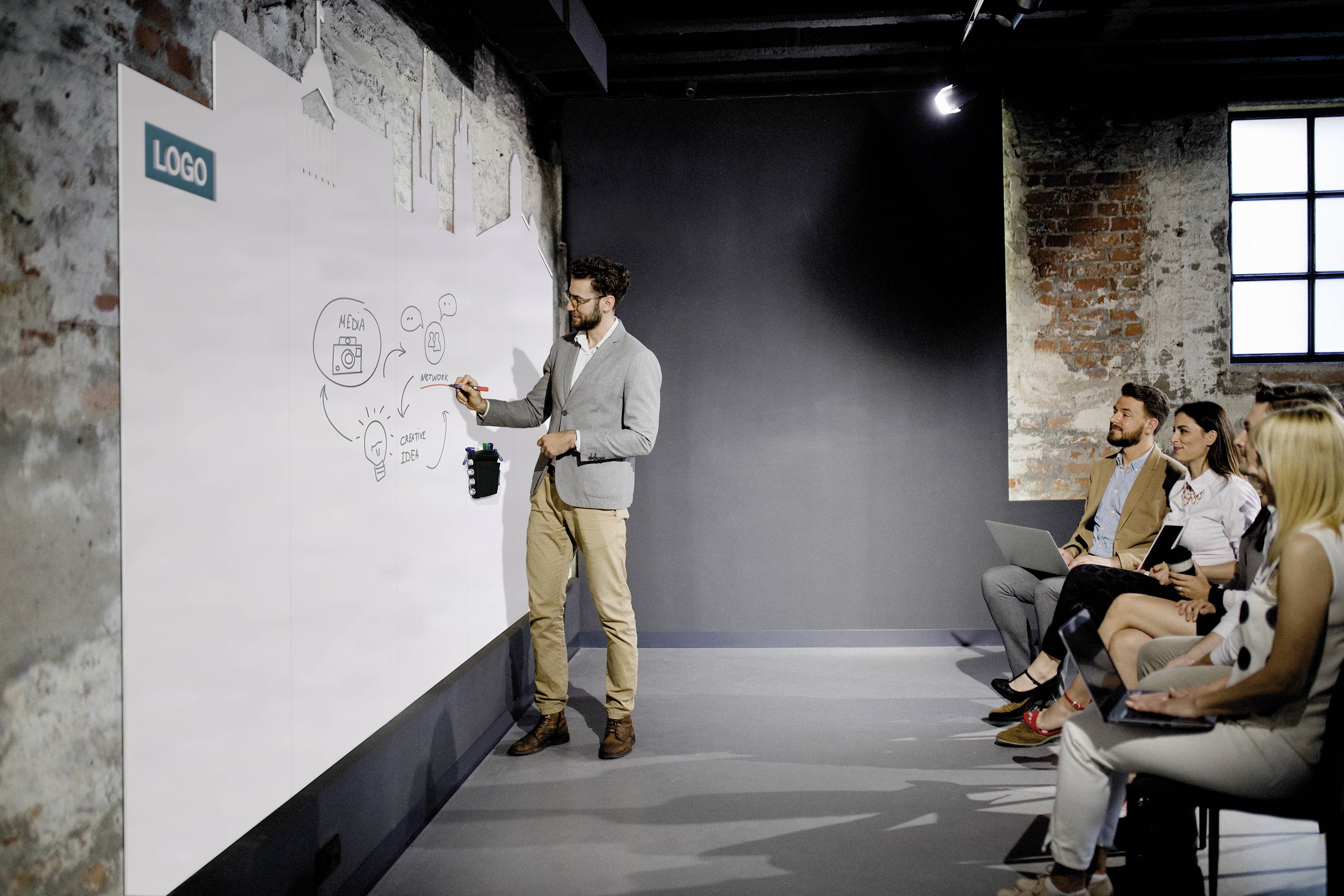 Mężczyzna w szarym swetrze stoi przed białą tablicą i wyjaśnia szkic. Kilka osób siedzi na krzesłach, słuchając go.