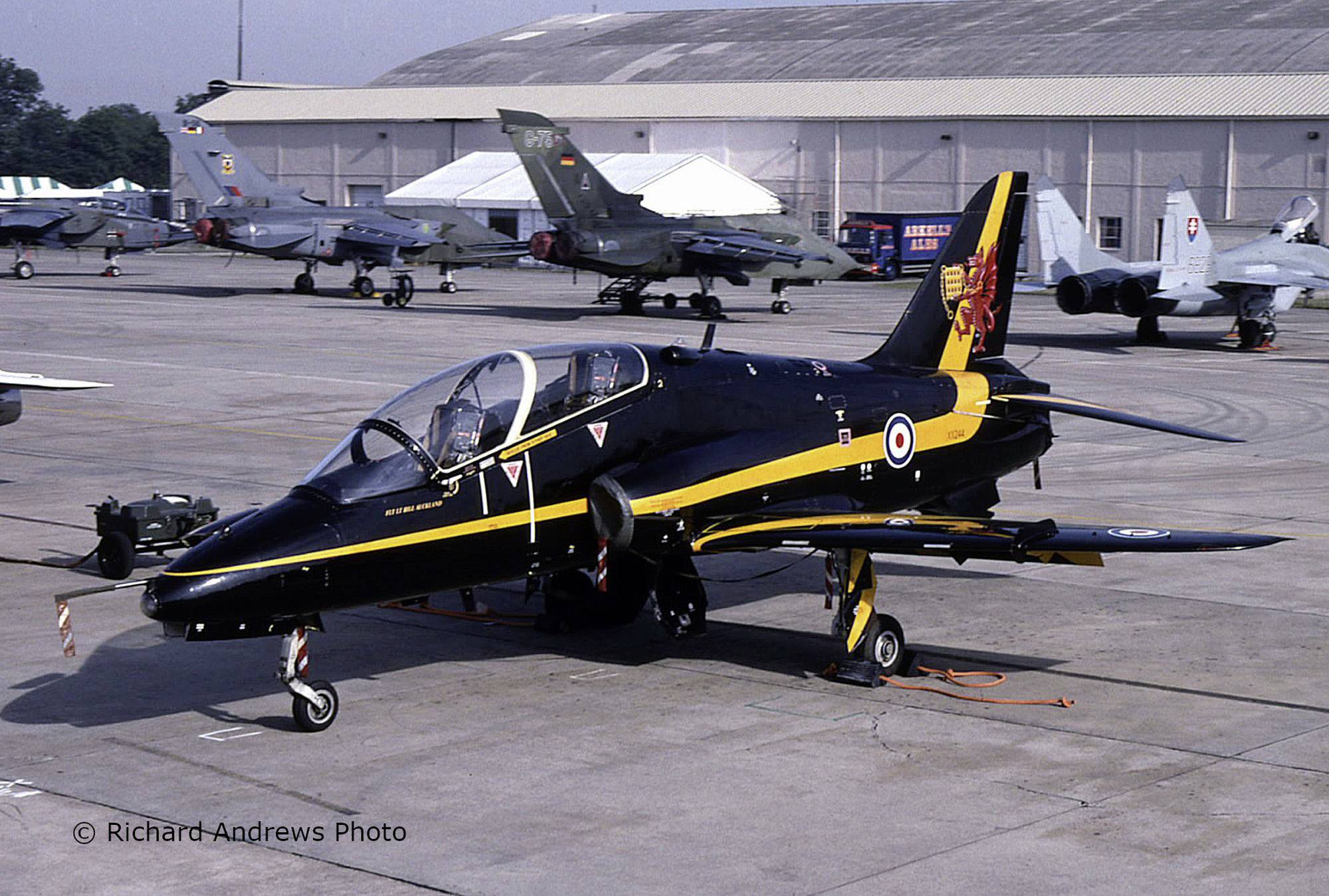 Czarno-żółty samolot akrobacyjny stoi na pasie startowym przed kilkoma wojskowymi myśliwcami w hangarze.