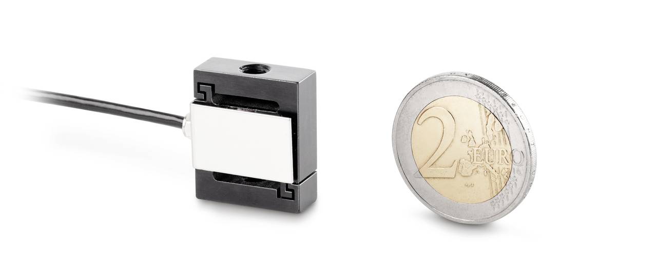 Mały, prostokątny czujnik magnetyczny obok monety 2-euro dla porównania rozmiaru. Czujnik jest czarny z metalowym rdzeniem.