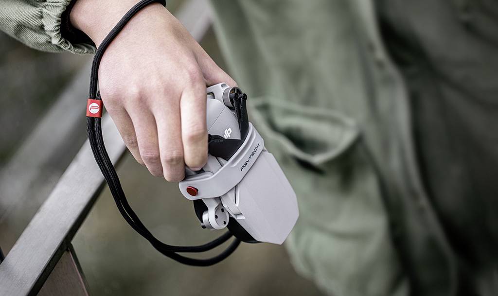 Dłoń trzyma kompaktowego drona z szarą obudową. Na tle widoczna jest zielona tkanina, prawdopodobnie fragment odzieży.