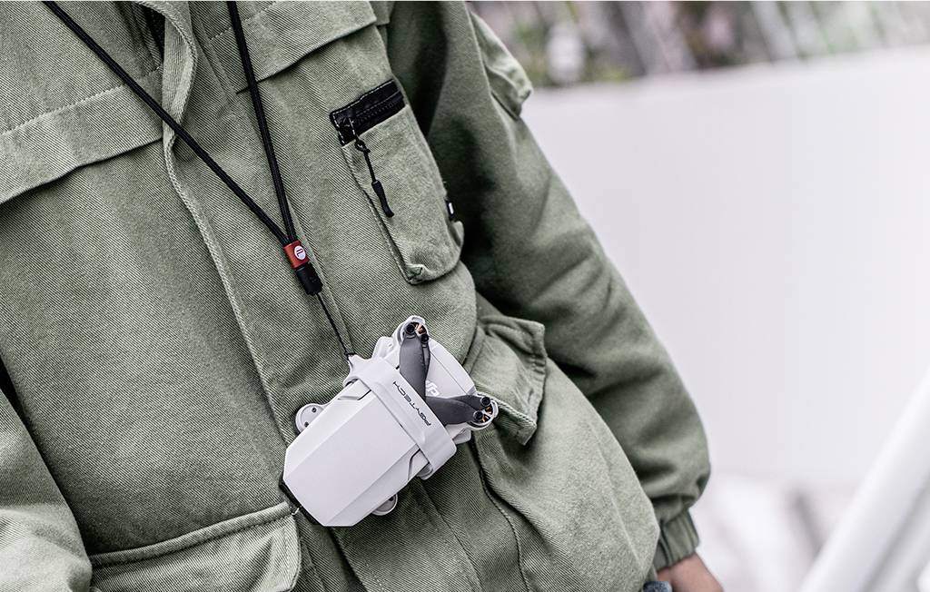 Osoba nosi zamkniętego drona zawieszonego na szyi, przymocowanego do paska. Osoba ma na sobie kurtkę w kolorze oliwkowym.