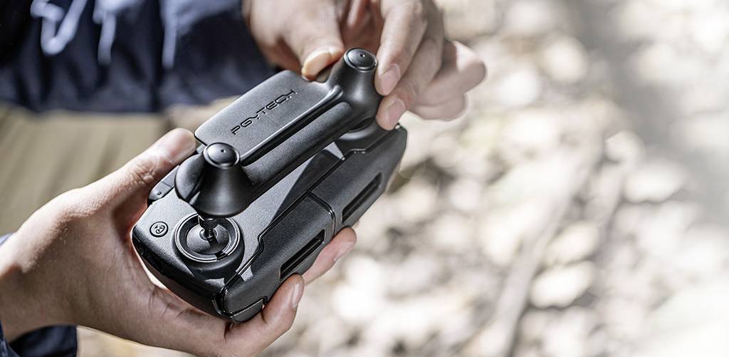 Ręce trzymają kompaktową, złożoną drona z nazwą marki na górnej powierzchni. Otoczenie jest lekko rozmyte i naturalne.