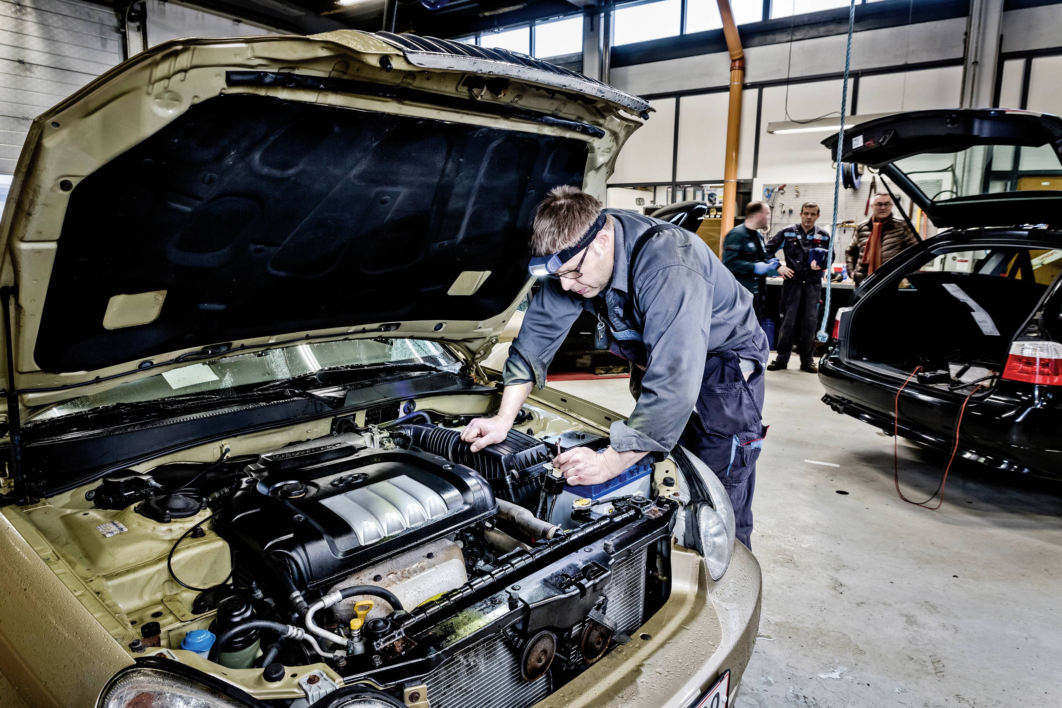 Mechanik naprawia samochód w warsztacie, w tle widać inne pojazdy i ludzi.