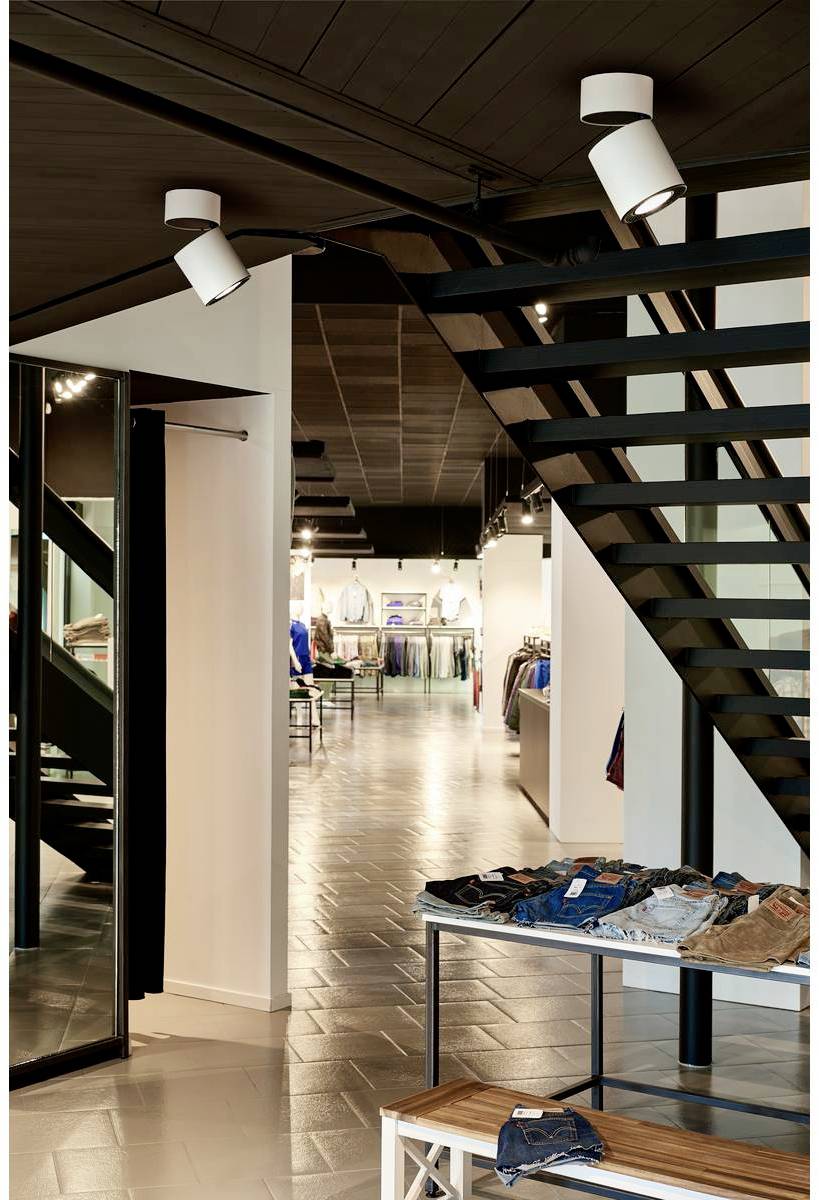 Nowoczesny sklep odzieżowy z czarnymi metalowymi schodami, stolikami prezentacyjnymi z jeansami i koszulami oraz dobrze oświetlonymi półkami.