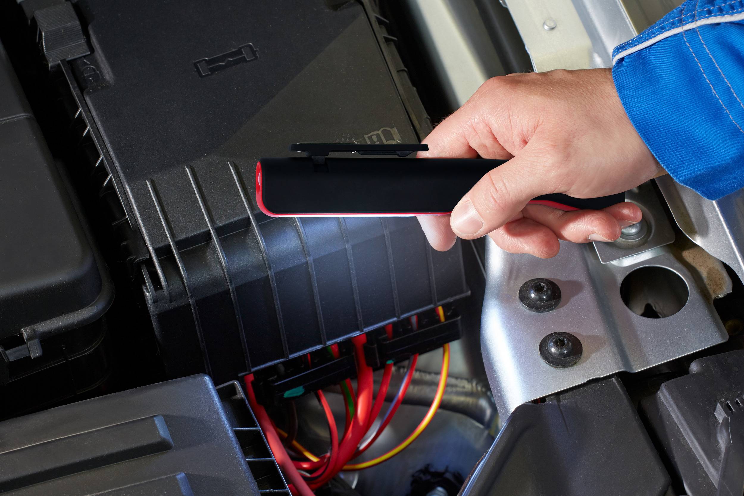 Osoba trzyma przenośny rozrusznik nad akumulatorem samochodu, gotowa do użycia. Kable i komponenty silnika są widoczne.