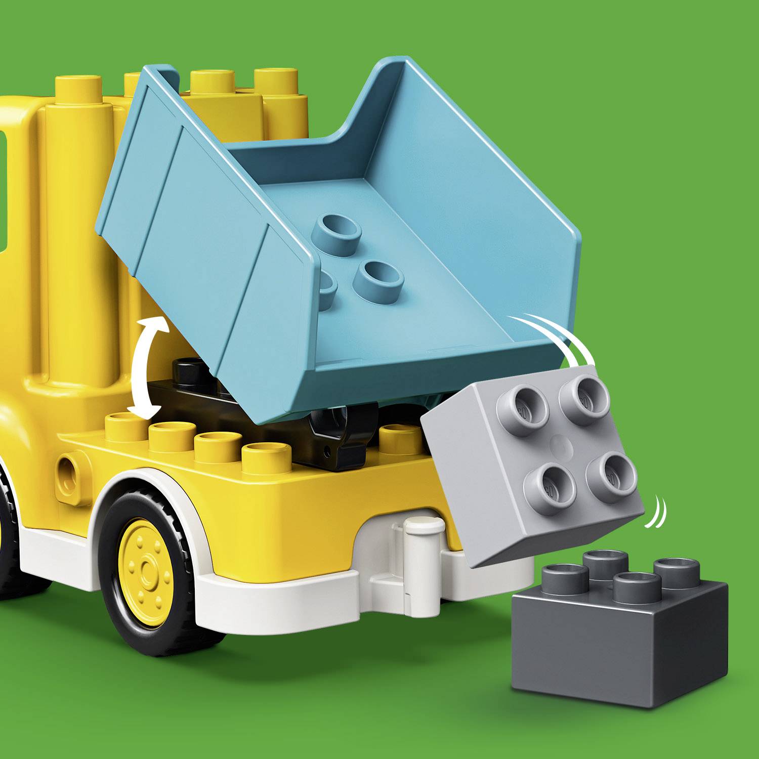 Żółta zabawkowa ciężarówka z klocków wywraca swoją niebieską skrzynię ładunkową, podczas gdy szary klocek wypada. Tło zielone.
