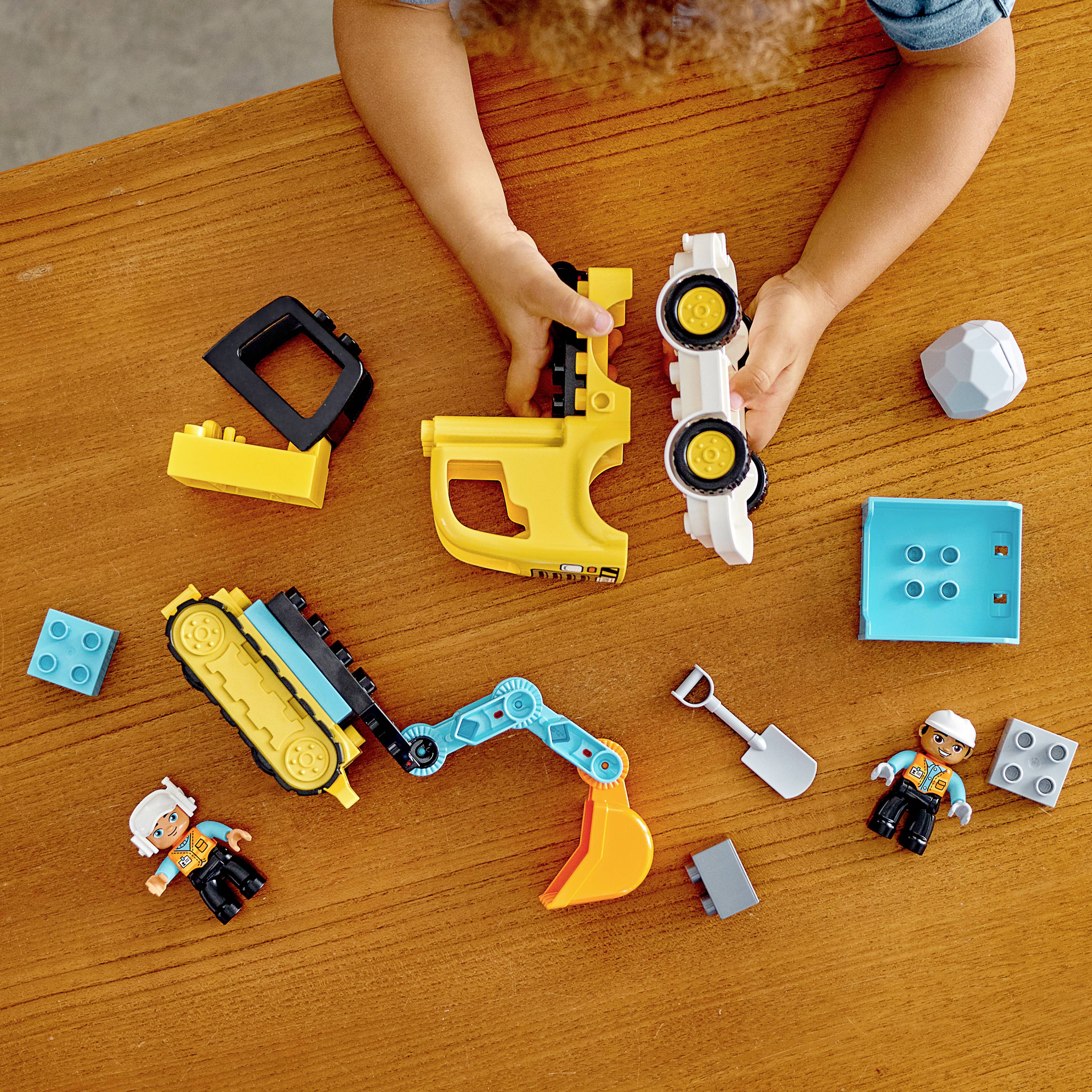 Dziecko bawi się kolorową zabawką na drewnianym stole, w tym klockami, figurkami i żółtą koparką.