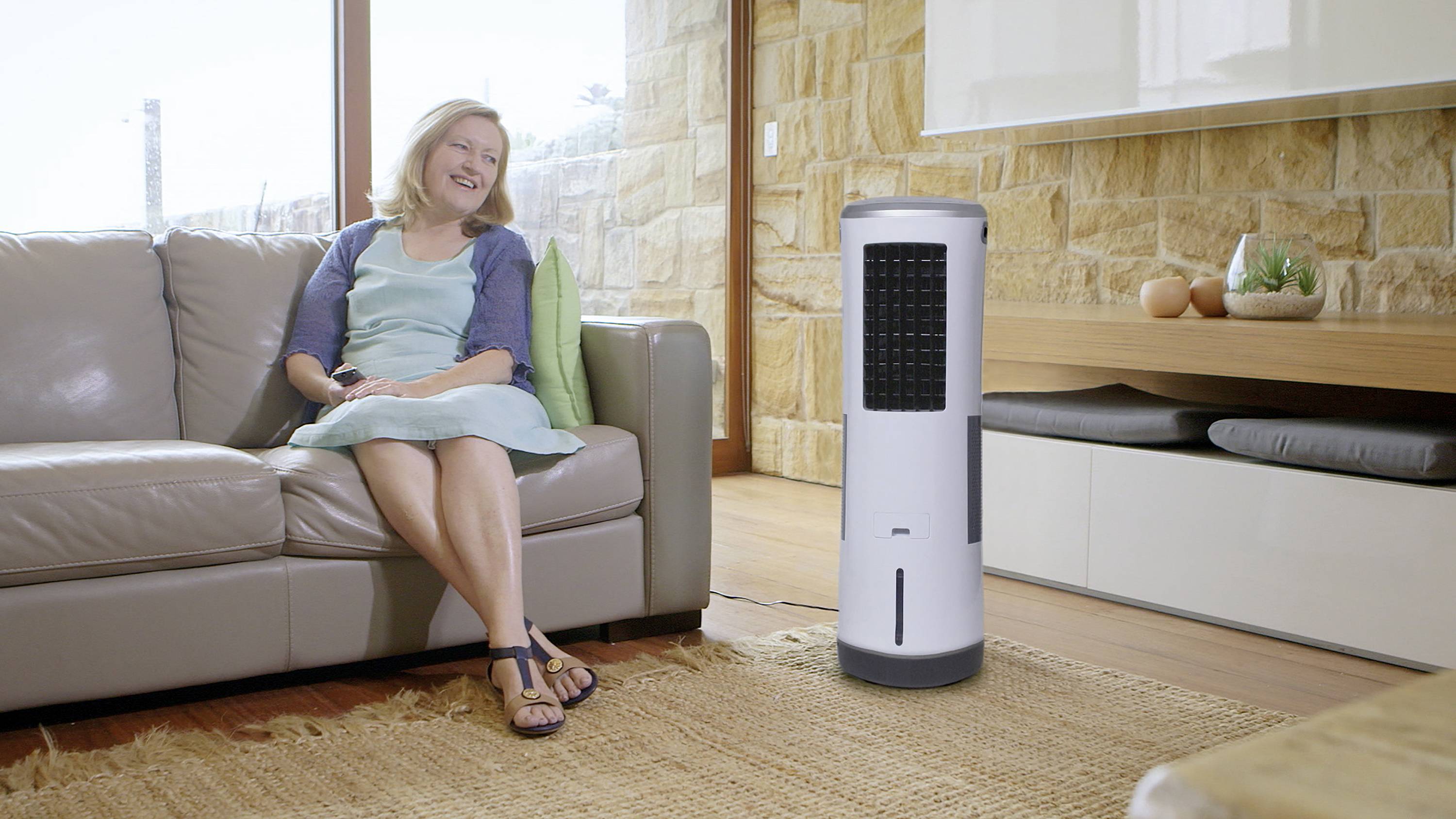 Kobieta siedzi uśmiechnięta na kanapie w salonie, obok niej na dywanie stoi przenośny klimatyzator.