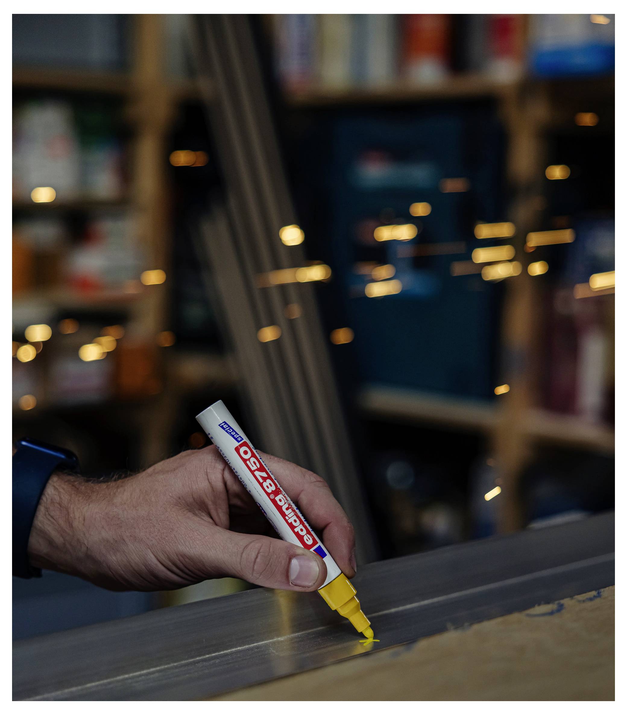 Ręka używa markera ze żółtym wierzchołkiem, aby zrobić znacznik na szarej powierzchni, z rozmazanymi półkami warsztatu w tle.