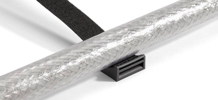 Zbliżenie czarnego plastikowego zacisku trzymającego pleciony kabel na białej powierzchni. Nad nim widoczny jest rzep.