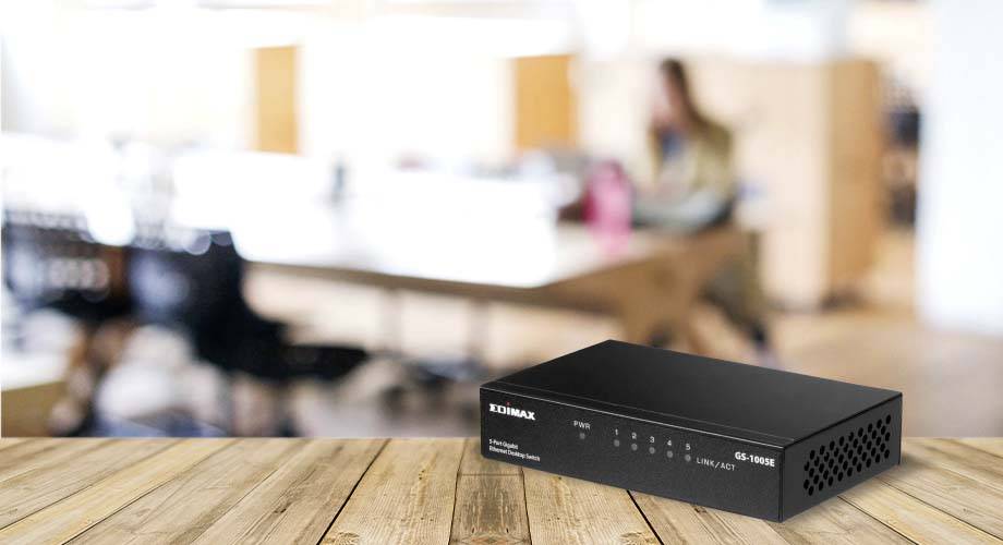 Przełącznik sieciowy stoi na drewnianym stole, w tle rozmyta przestrzeń biurowa.