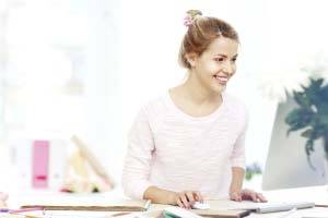 Uśmiechnięta kobieta pracuje przy komputerze, otoczona materiałami biurowymi. Ma na sobie jasną górę i włosy związane w górze.