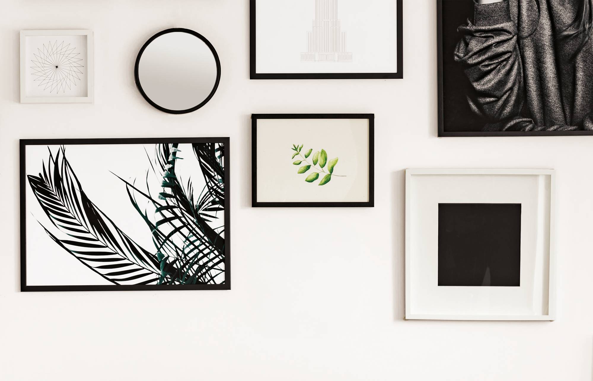 Galeria ścienna z oprawionymi pracami sztuki i grafikami, obejmująca czarno-białe projekty botaniczne, okrągłe lustro i obrazy abstrakcyjne na białej ścianie.