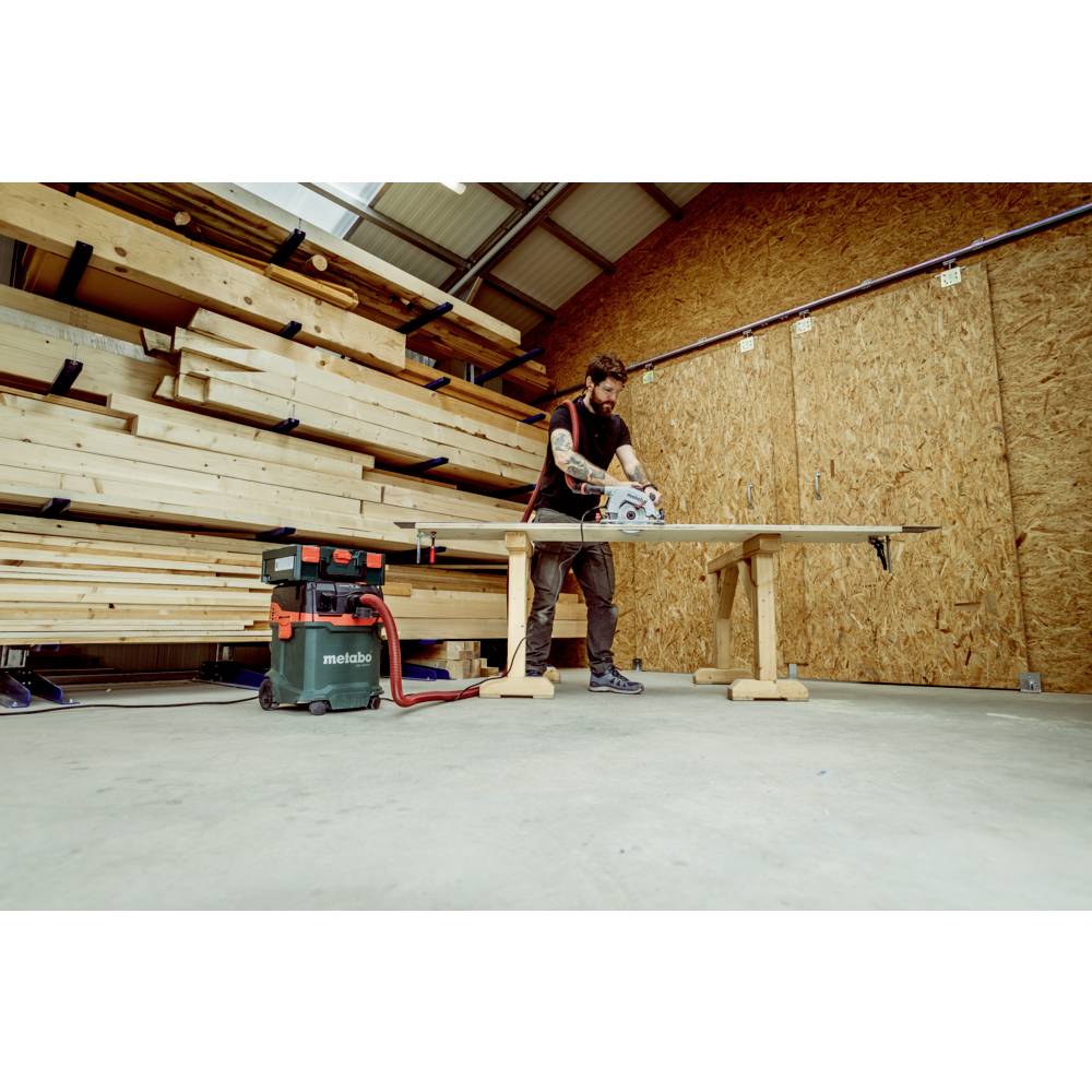 Mężczyzna w warsztacie tnie drewnianą deskę na stole za pomocą piły tarczowej. W tle widoczne są stosy drewna.