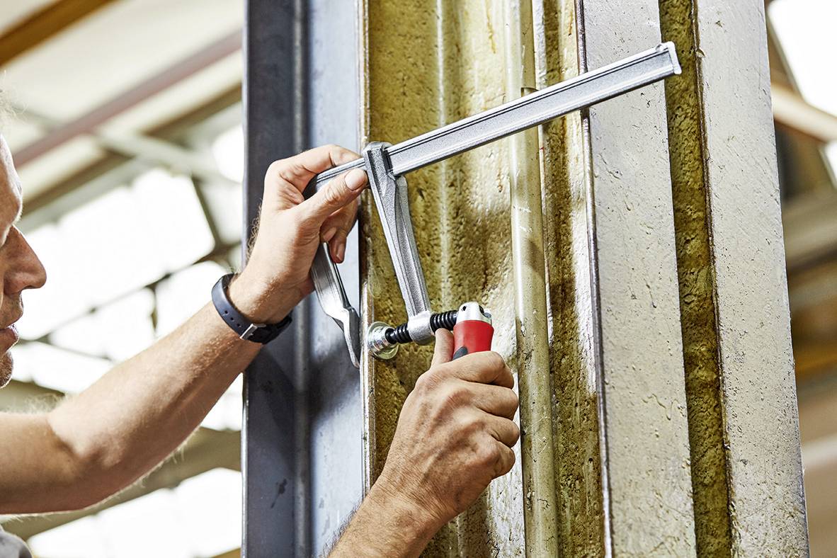 Osoba używa zacisku śrubowego, aby docisnąć dwie metalowe powierzchnie. Ręce obracają rękojeść, dokręcając ją. W tle widoczne jest środowisko warsztatowe.
