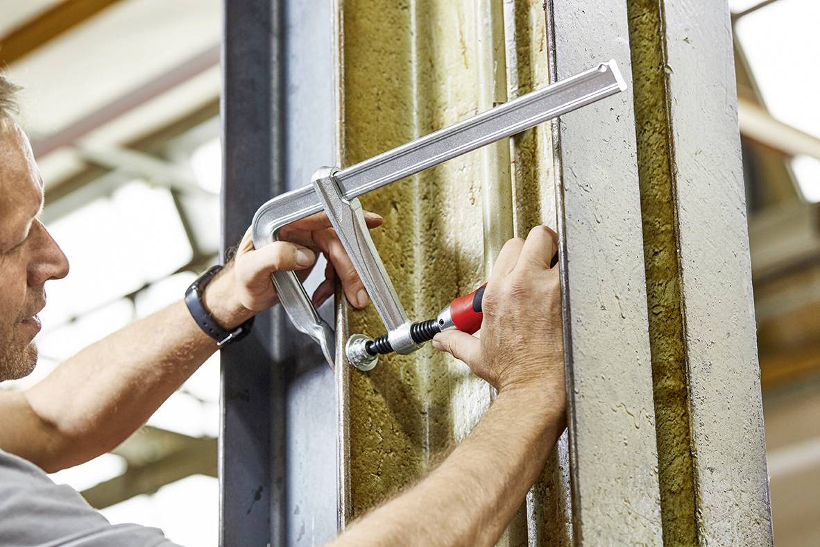 Mężczyzna mocuje element metalowy zaciskiem ślusarskim do belki. Używa śrubokręta, aby zabezpieczyć zacisk.