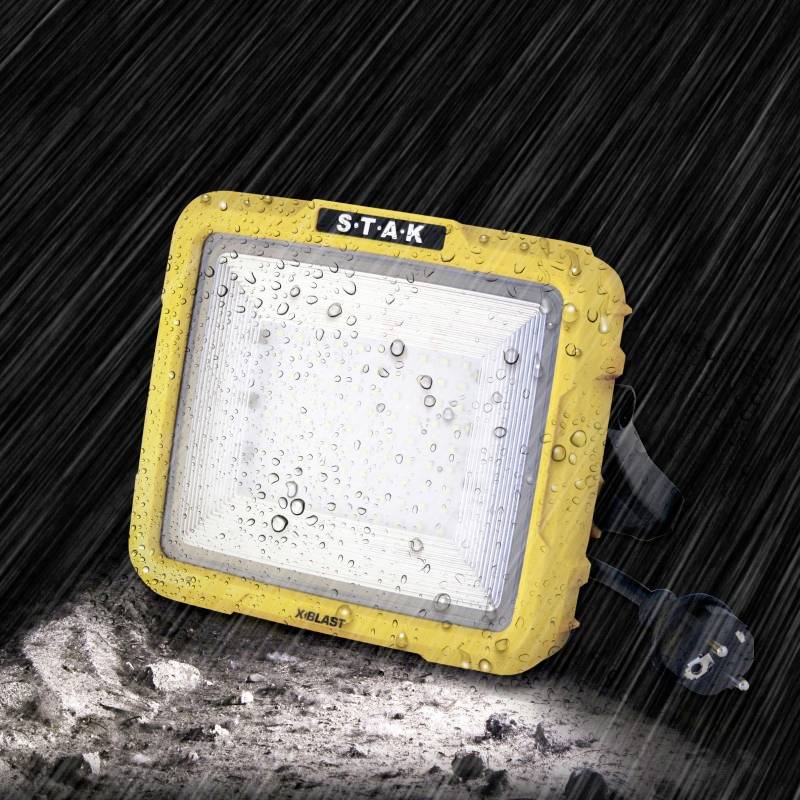 'Mokry, żółty reflektor budowlany w deszczu na wilgotnej ziemi, z wtyczką z boku.'