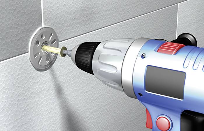 Wiertarka wwierca otwór w szarej płytce ceramicznej. Wiertło jest srebrno-niebieskie z czerwonym włącznikiem, a wokół sypie się pył.