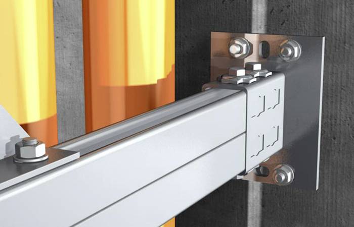 Metalowy nośnik zamocowany na ścianie; pomarańczowe rury biegną równolegle. Stabilne podparcie, industrialne otoczenie.