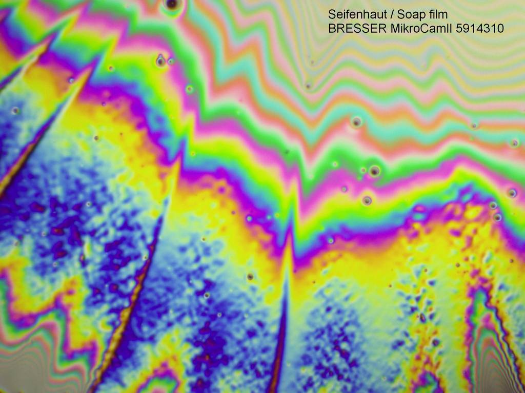 Zbliżenie powierzchni błony mydlanej ukazuje kolorowe, faliste wzory w barwach tęczy. W górnym prawym rogu widnieje napis 'Seifenhaut / Soap film'.