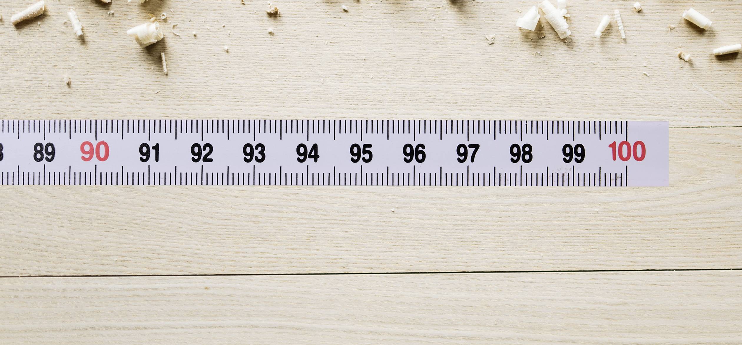Miarka krawiecka na drewnianym stole pokazuje centymetry od 90 do 100. Wokół miarki rozrzucone są wióry drewniane.