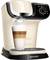 Ekspres do kawy z logo Tassimo przygotowuje latte macchiato w przezroczystej szklance. Para unosi się nad napojem.