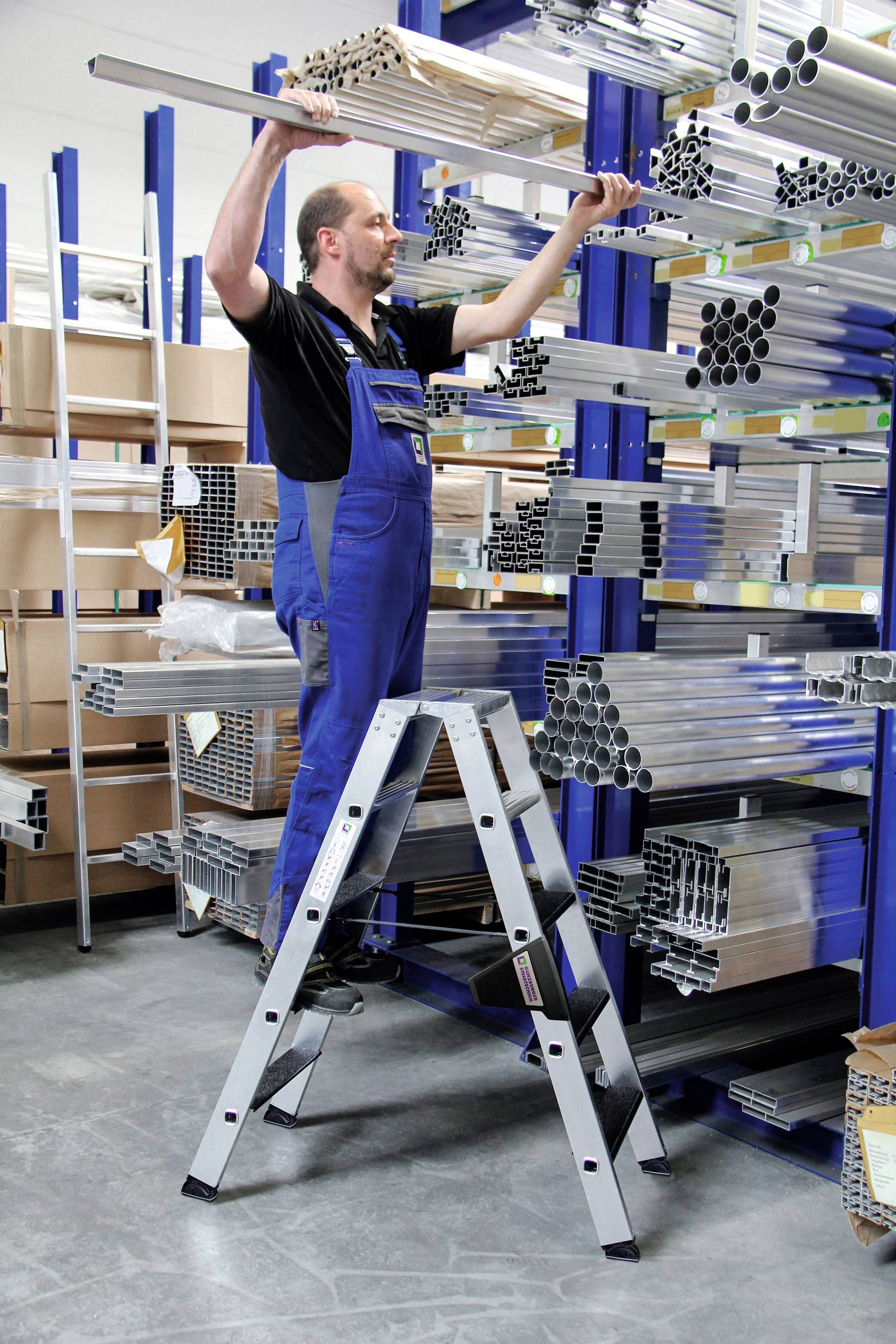 Mężczyzna w niebieskim ubraniu roboczym stoi na drabinie w magazynie i porządkuje metalowe rury na półkach.