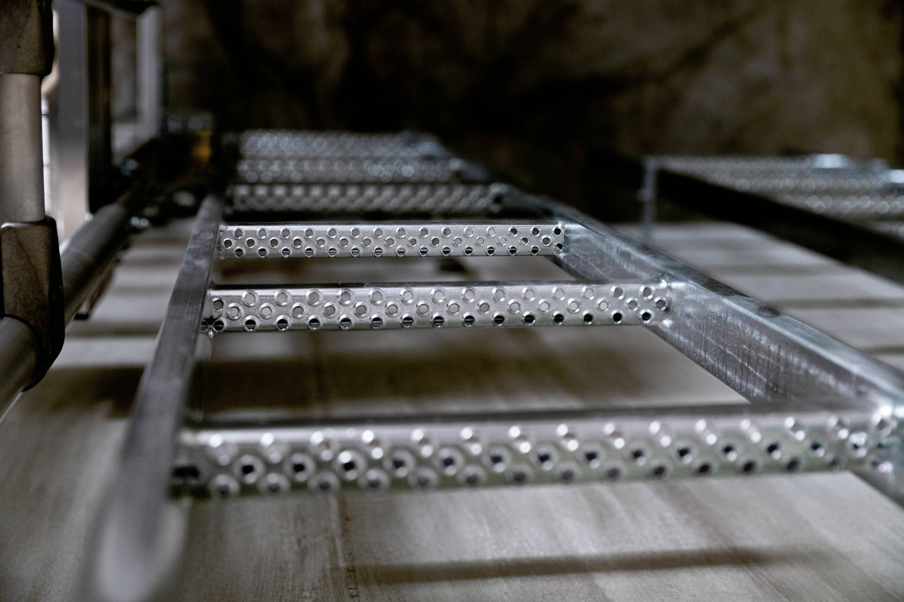 Metalowa drabina prowadząca skośnie w górę. Fokus na szczeblach, ukazujących szczegóły struktury i grubości materiału. Rozmyte tło.