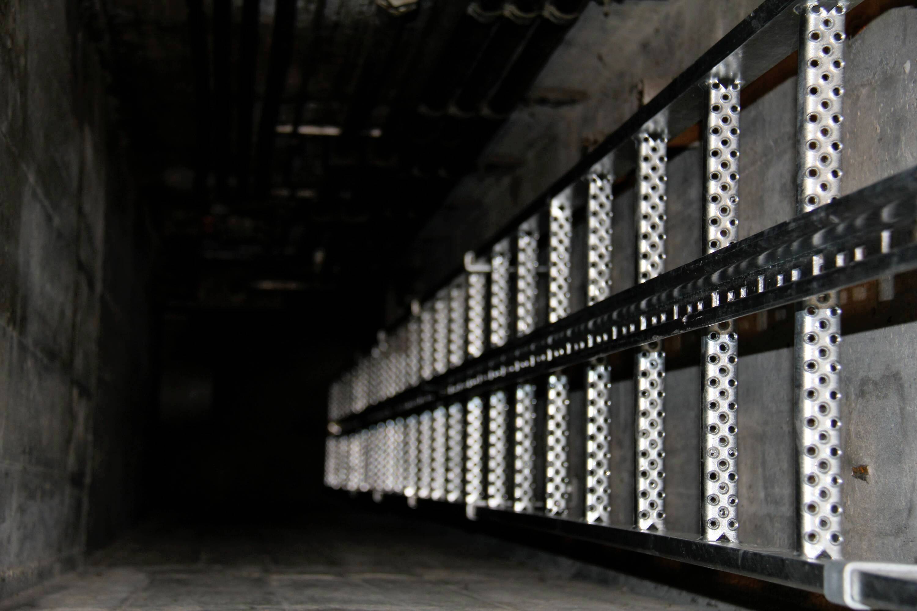 Metalowa drabina prowadzi pionowo w ciemny szyb. Na górze widoczne są rury i kable.