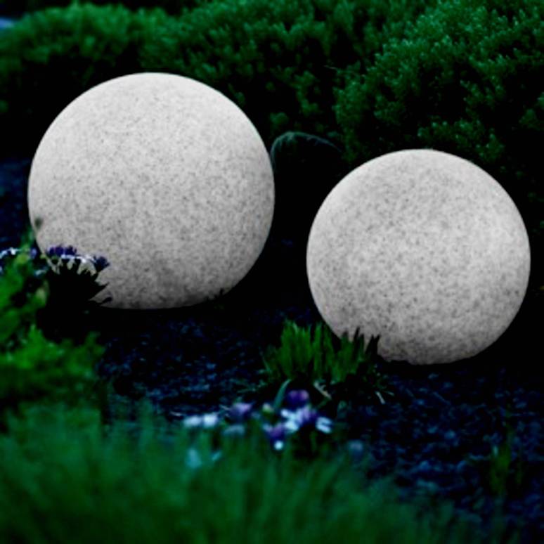 Dwie szare kamienne kule o różnej wielkości leżą na ciemnej powierzchni, otoczone zieloną roślinnością.