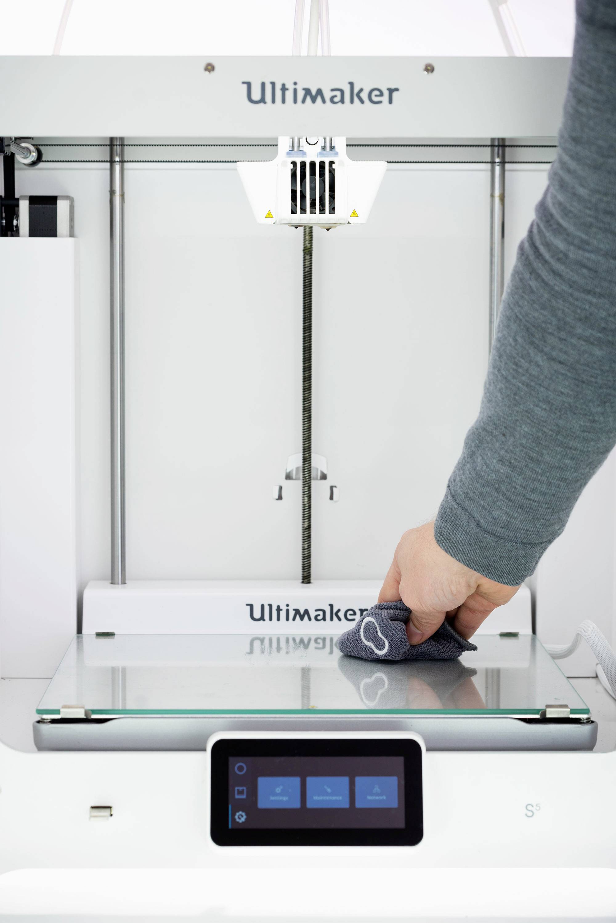 Drukarka 3D jest czyszczona kawałkiem tkaniny. Fokus jest skierowany na dłoń, która przesuwa szmatkę po platformie drukarki.