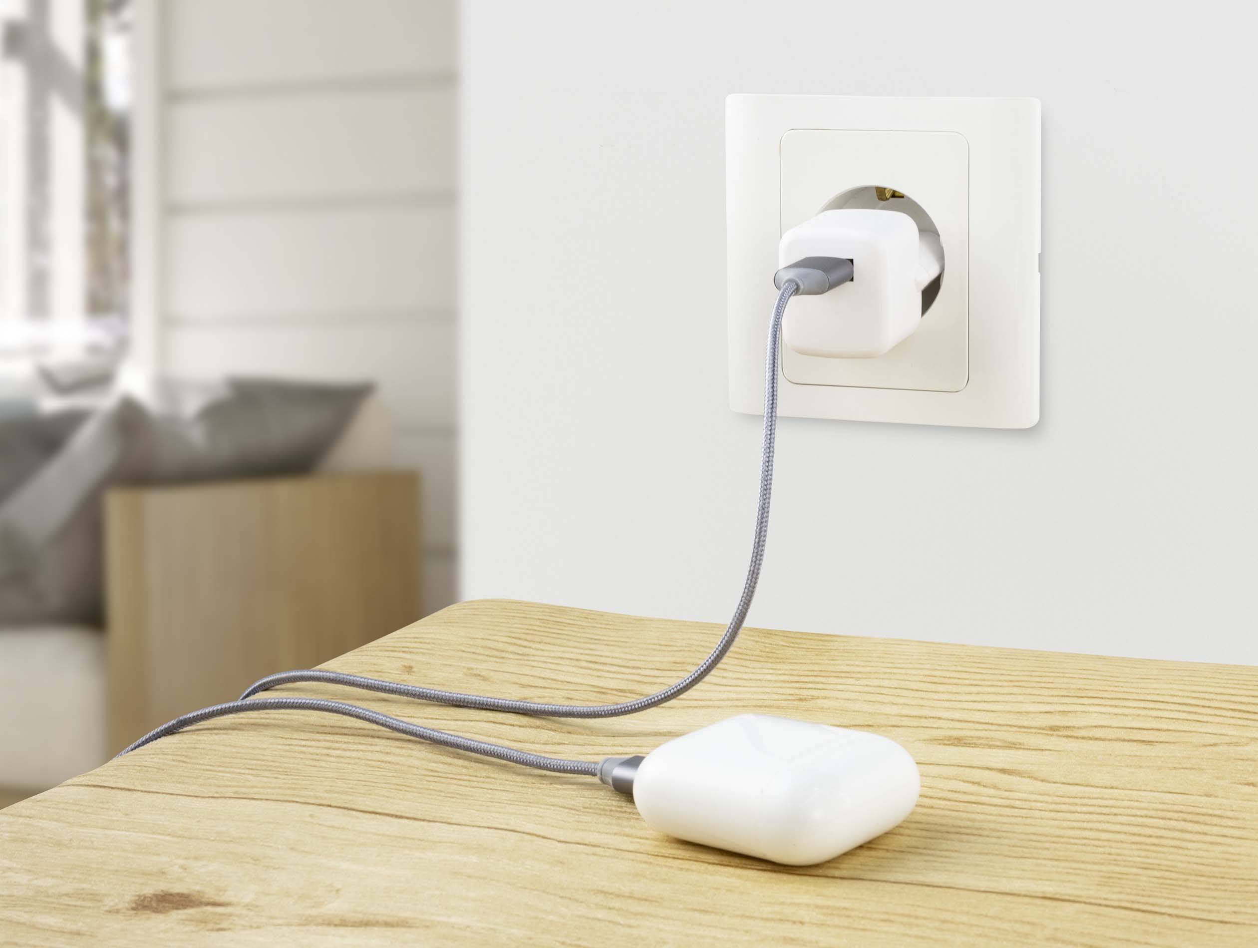 Białą ładowarkę włożono do gniazdka ściennego, a etui bezprzewodowych słuchawek leży na drewnianym stole.