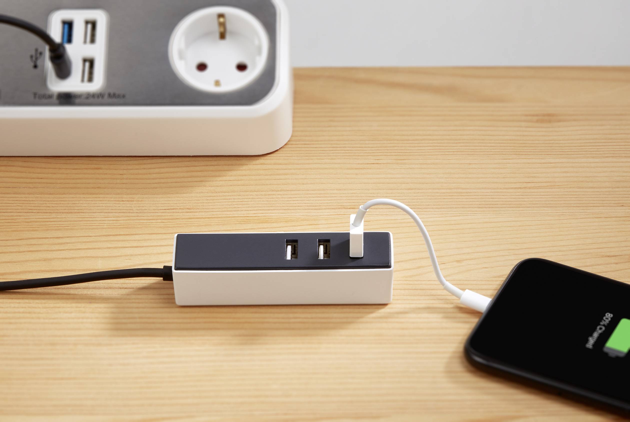 Telefon komórkowe jest podłączone przez kabel USB do adaptera, który leży na drewnianym stole obok listwy zasilającej.