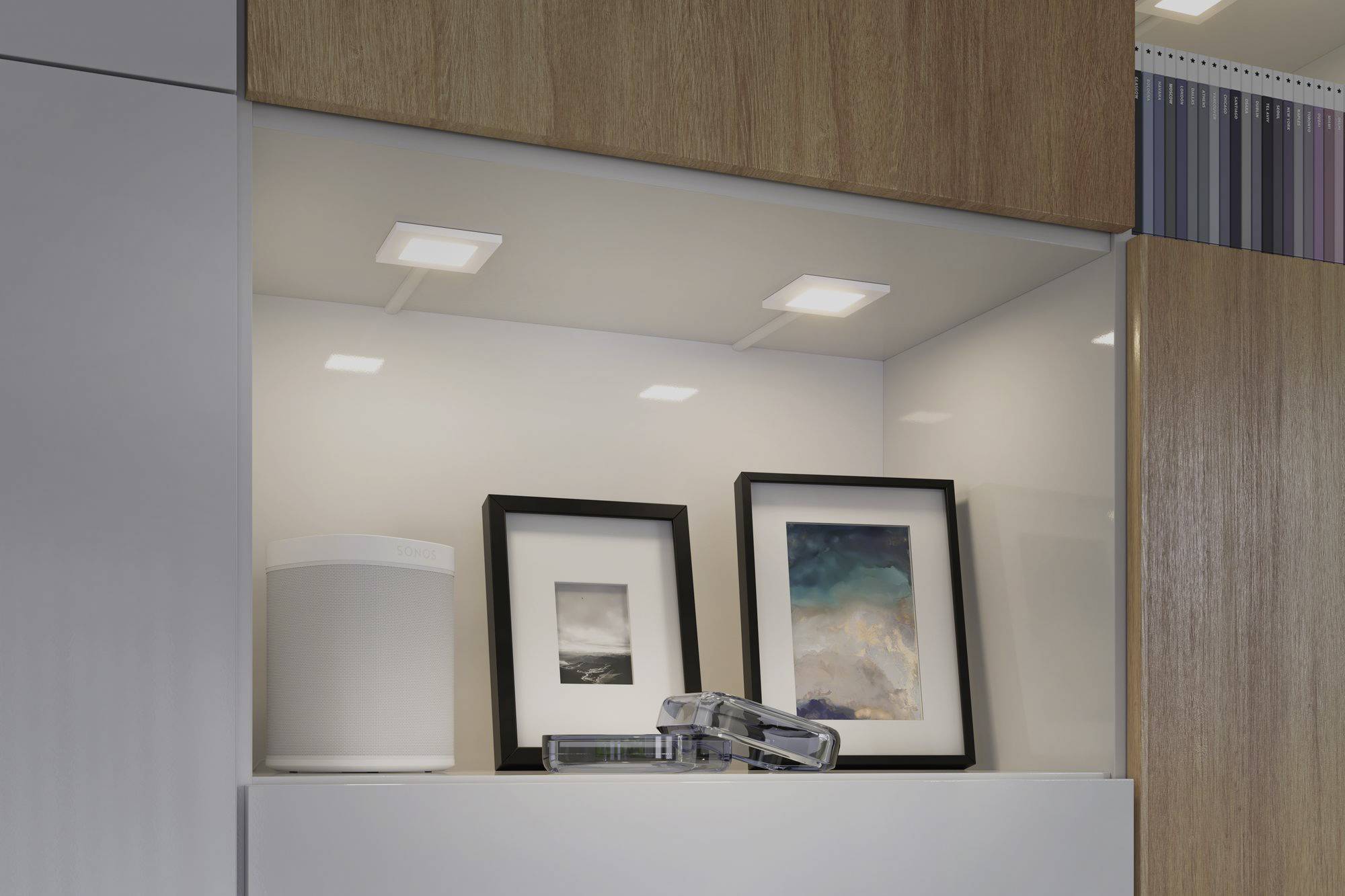 Półka z dwoma oprawionymi obrazami i białym pudełkiem. Oświetlona od góry, widoczne elementy drewniane i białe.