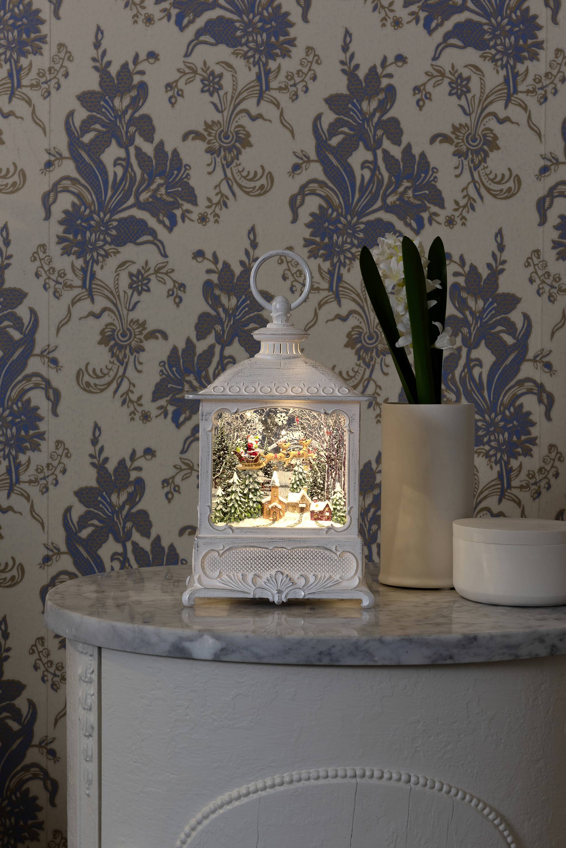 Biała latarnia na stole ukazuje świąteczną scenę z bałwankiem i lasem, oświetloną w nostalgicznym stylu.