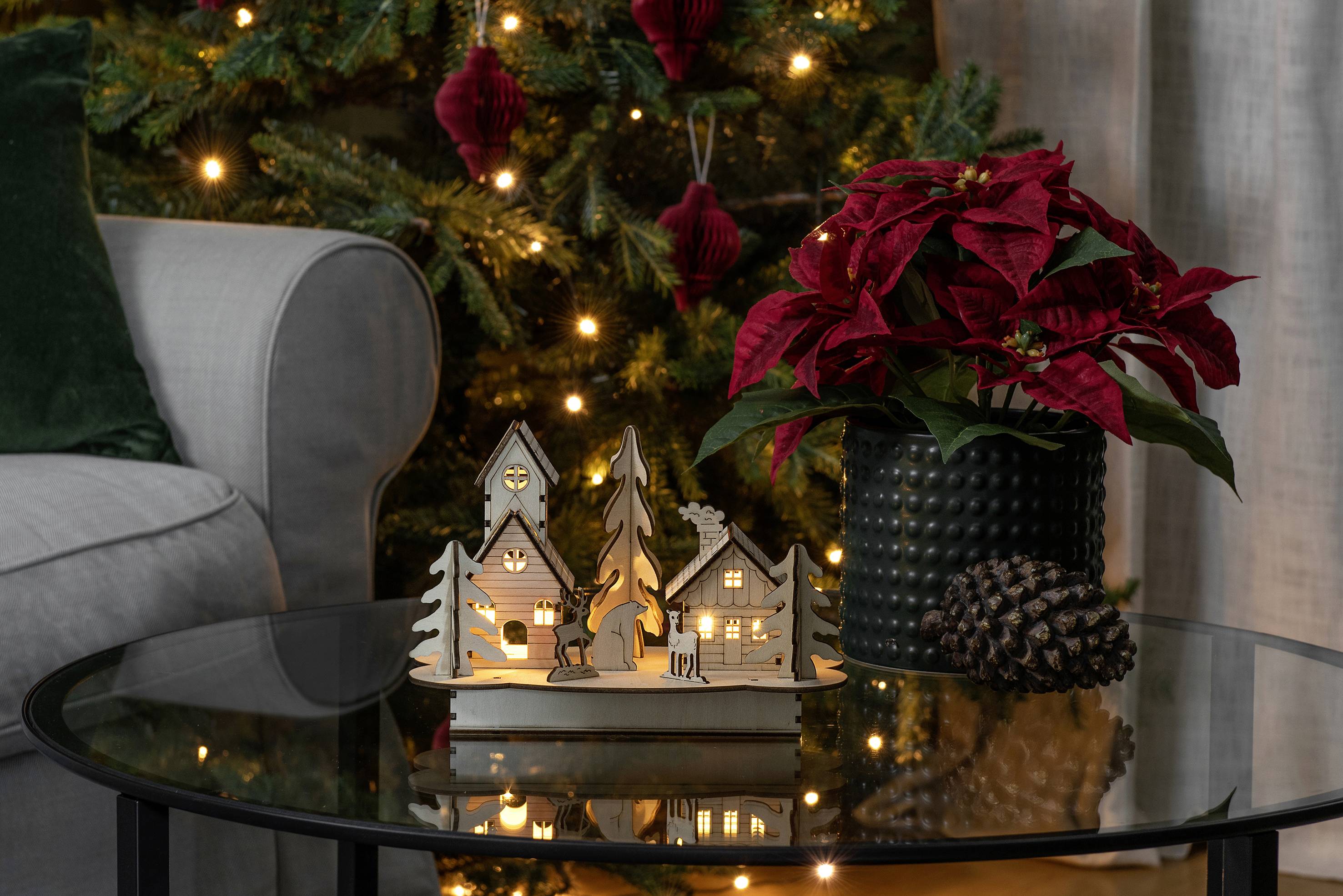 Udekorowany świąteczny stół z dekoracją z drewnianych domków i podświetloną choinką w tle. Obok stoi doniczka z czerwonymi poinsecjami.